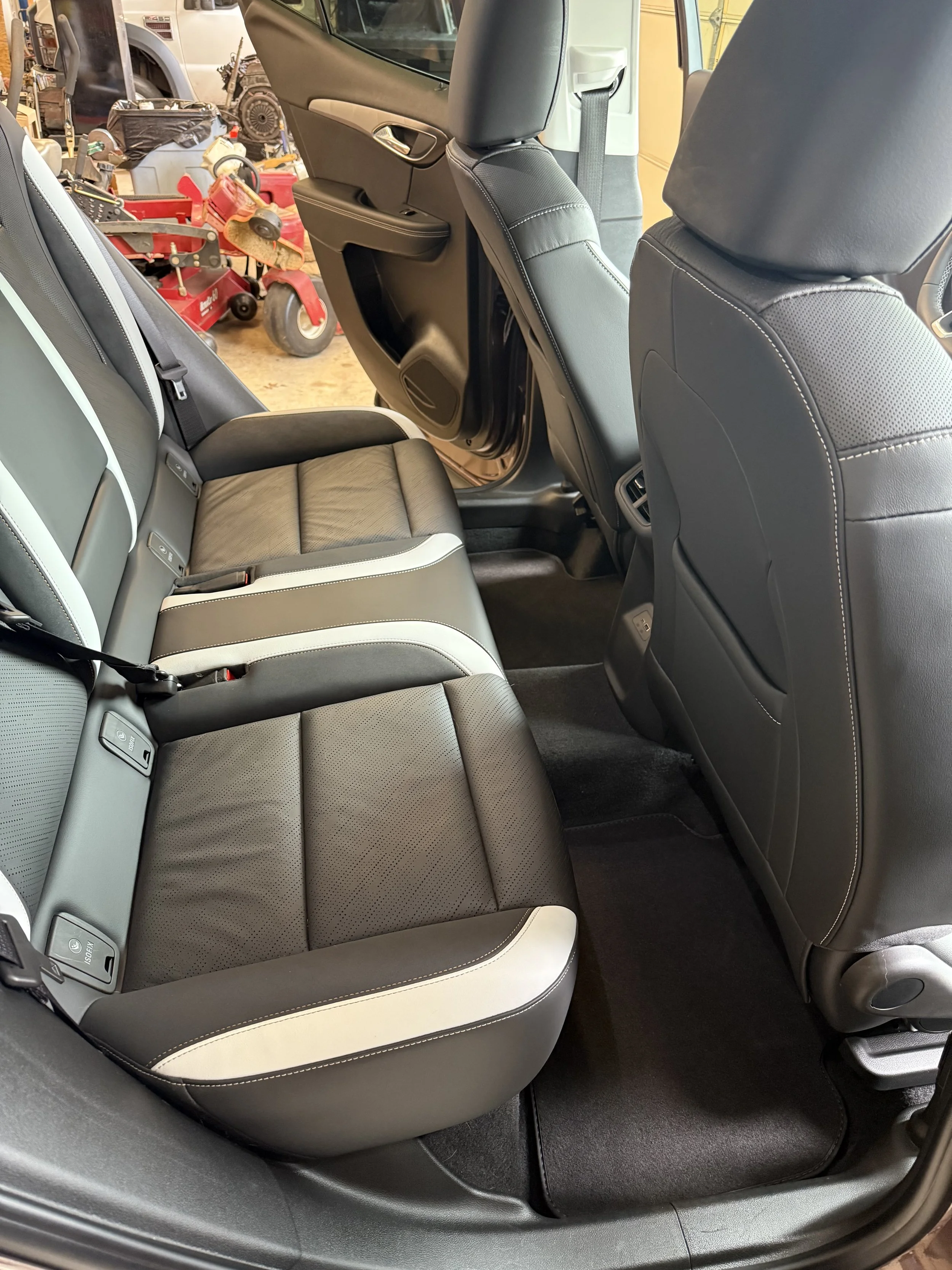 The backseat interior of a vehicle showing black and gray leather seats with white accents, grey floor mats, and the back of the front seats. A garage with tools and equipment is visible in the background.
