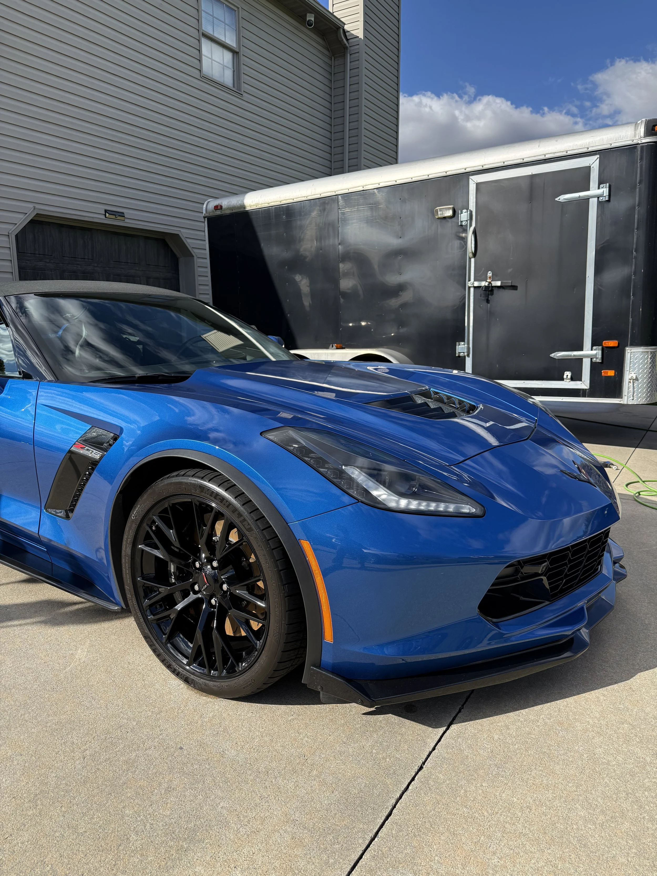 A blue Chevrolet Corvette sports car parked on a driveway next to a black trailer with a closed door and silver locks, in front of a beige house with a garage and a window, under a partly cloudy blue sky.