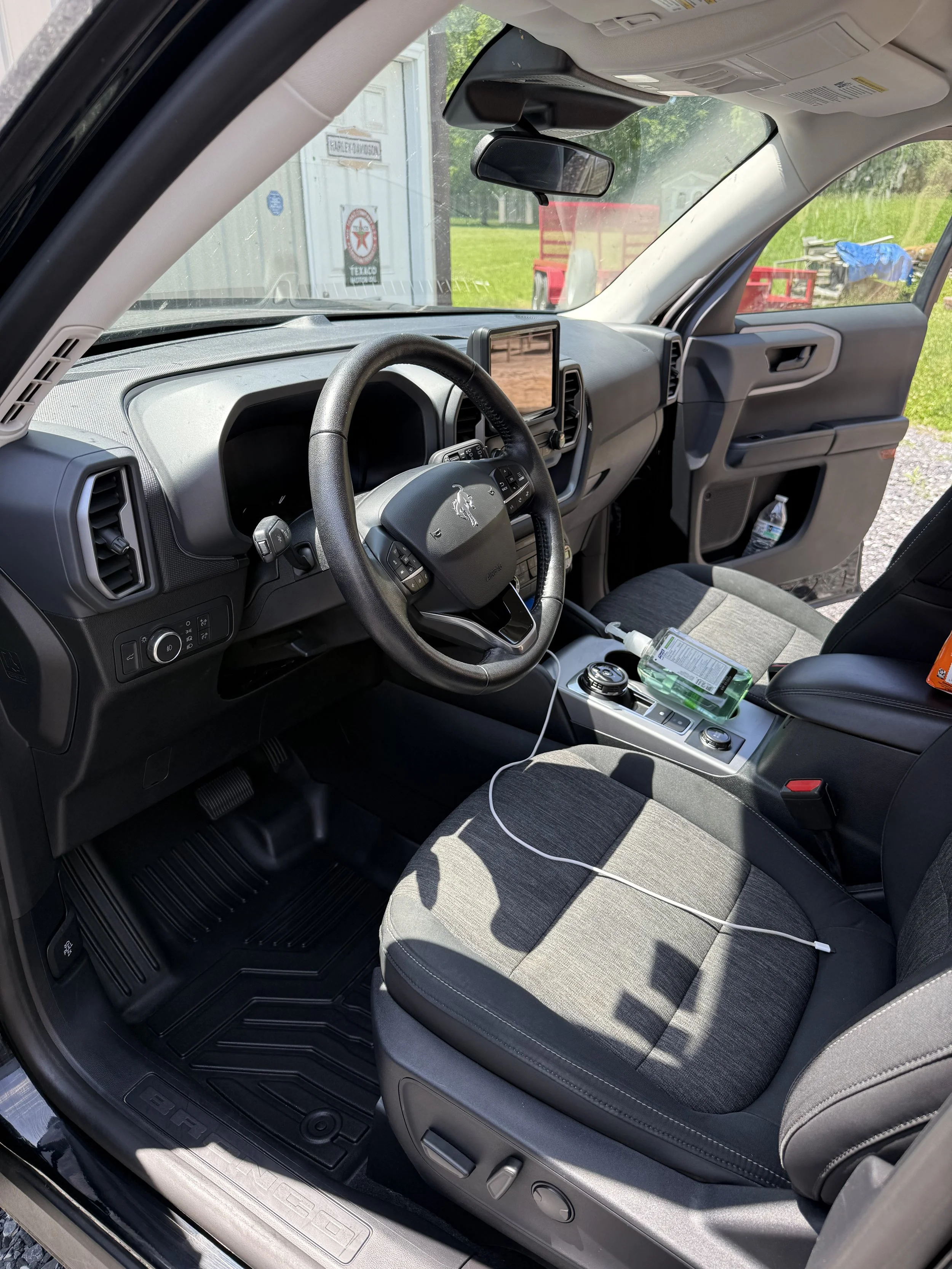 Interior of a vehicle, showing the driver's seat, steering wheel, dashboard, and center console with a phone charger, hand sanitizer, and other items. The door is open, revealing the outside surroundings.
