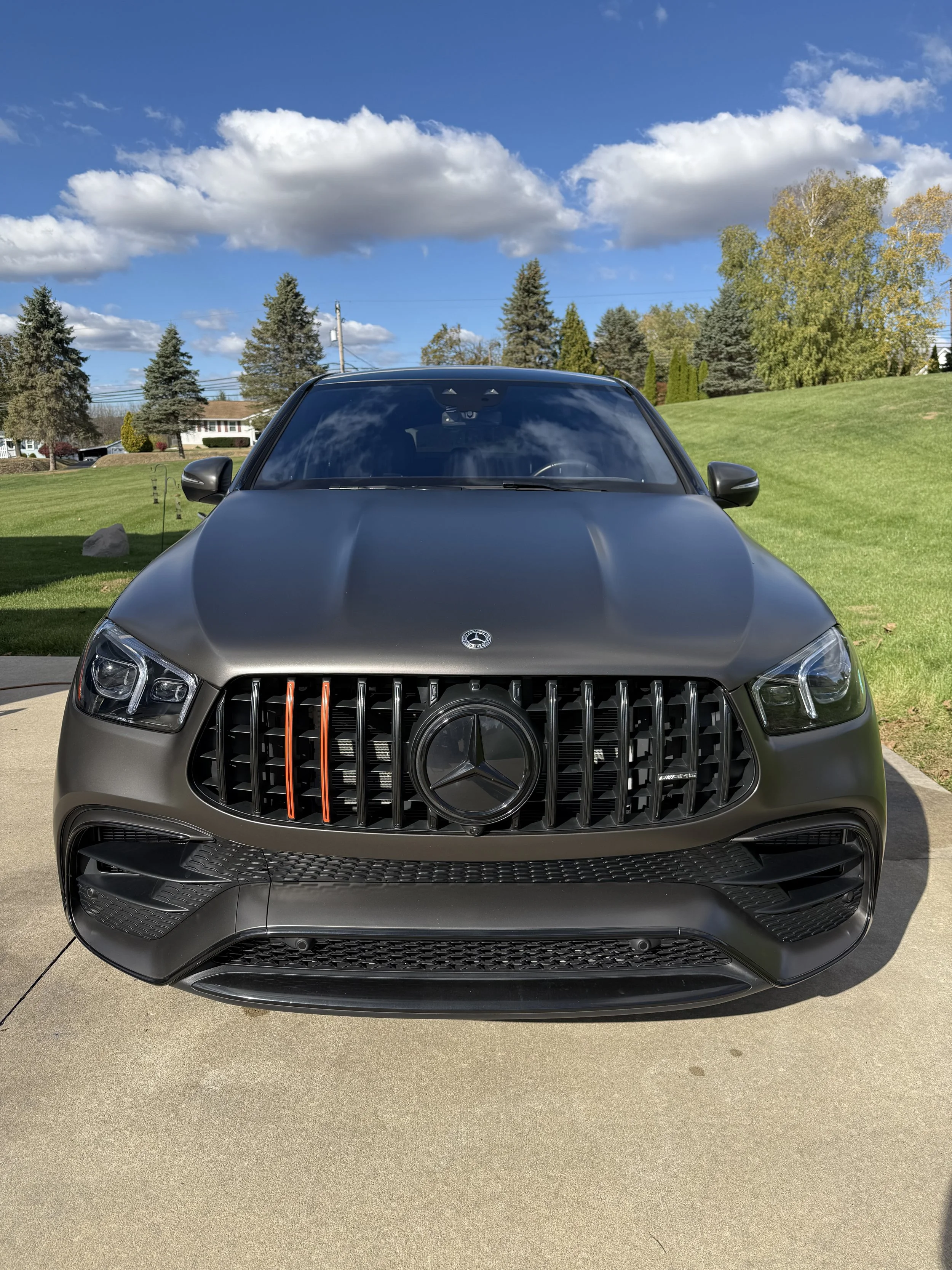 Front view of a matte black Mercedes-Benz vehicle parked on a driveway with green grass, trees, and blue sky with clouds in the background.