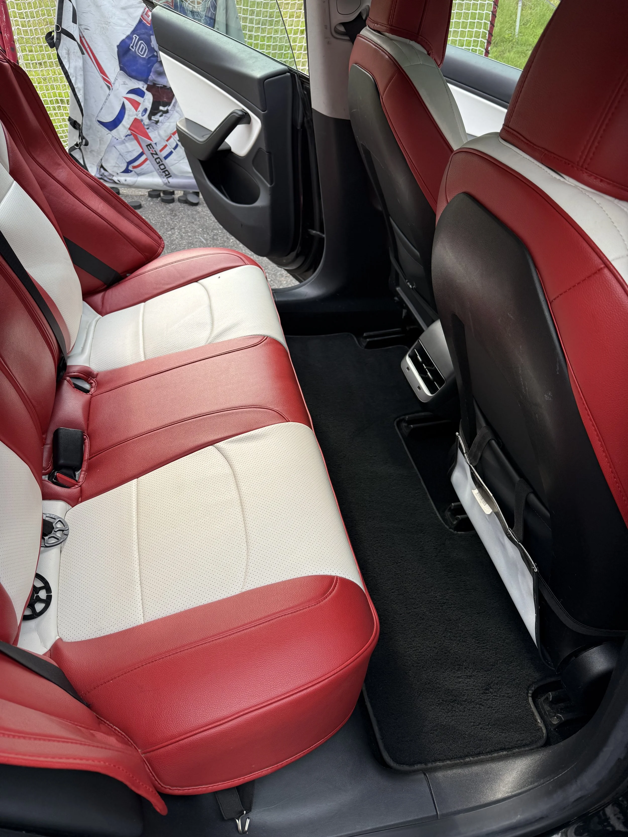Rear passenger seats of a vehicle with red and white leather upholstery, black floor mat, and back of front seats with black backings.
