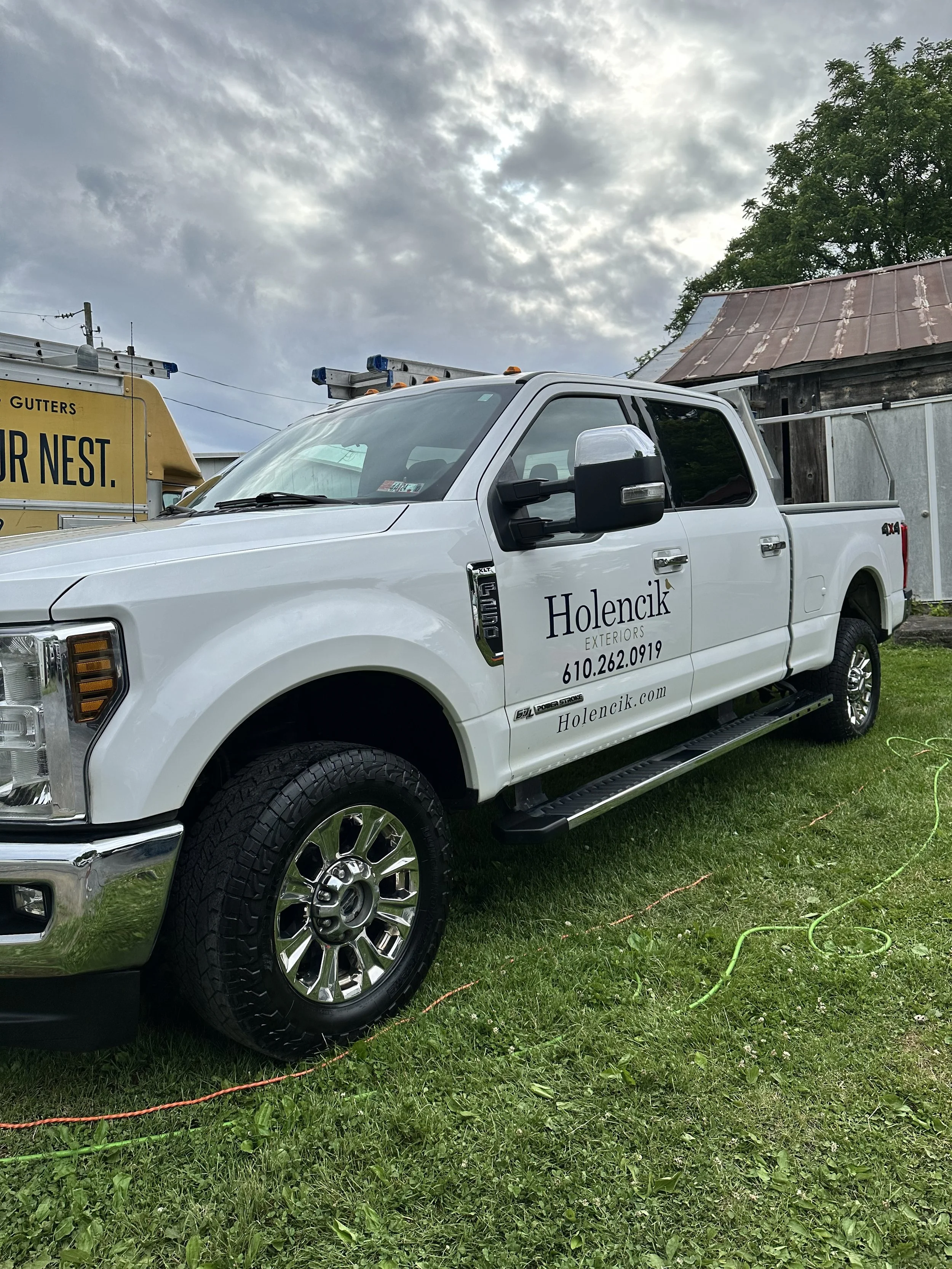 White pickup truck with black side mirrors and company's name 'Holencik Exteriors' on the door, parked on grass with overcast sky behind it.