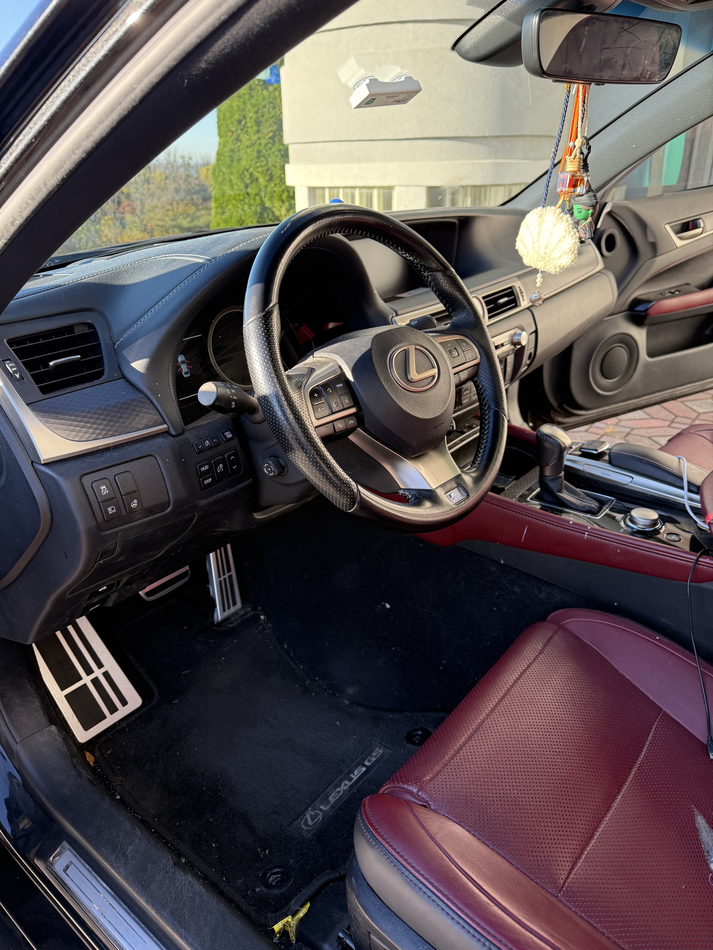 Interior of a Lexus car showing the steering wheel, dashboard, gear shift, and red leather seat with hanging decorations from the rearview mirror.