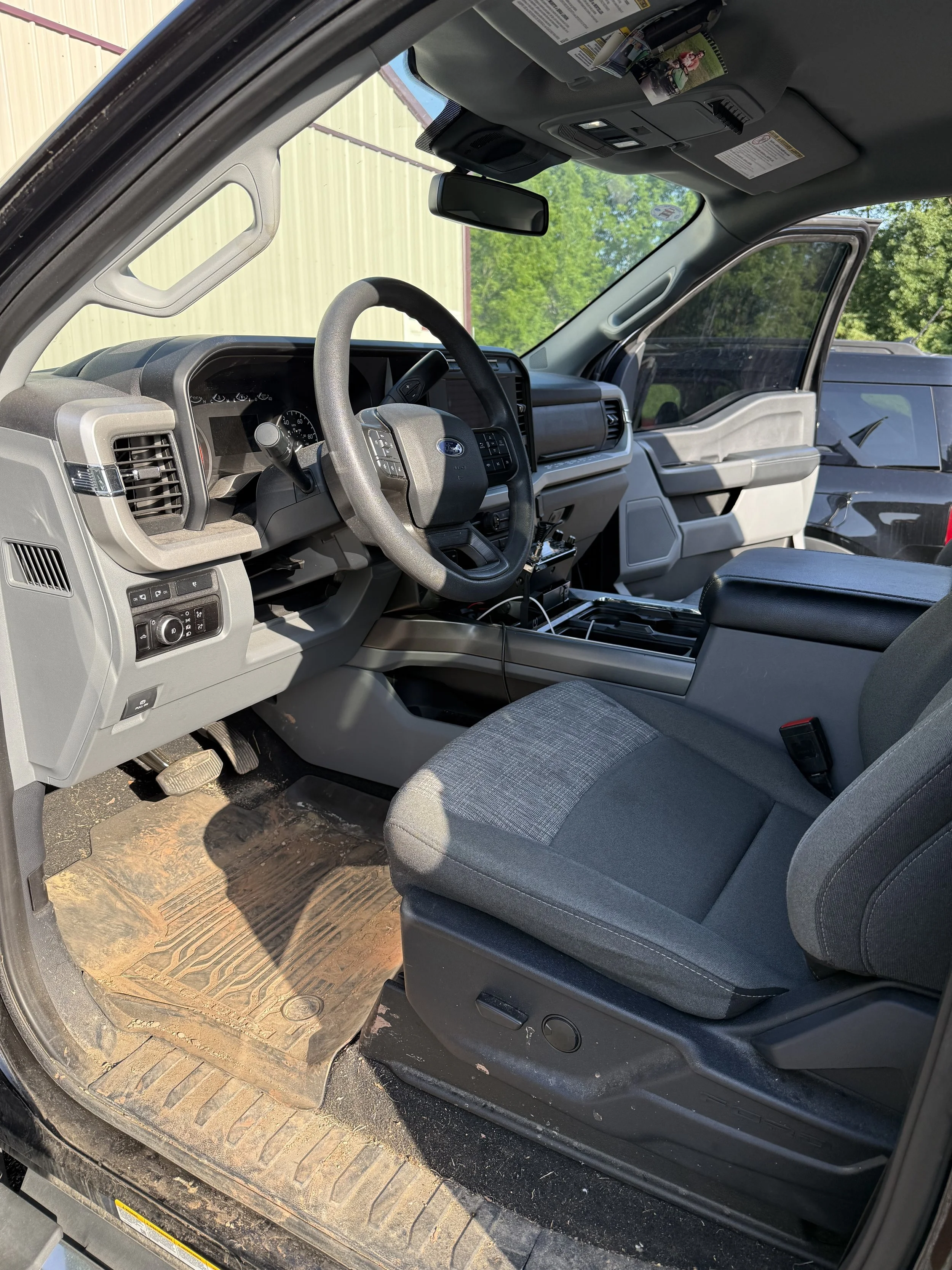 Interior of a pickup truck's front cabin, showing a gray fabric driver's seat, dashboard, steering wheel, and gear shift area with a dirt-covered rubber floor mat, and an external view of other vehicles and green trees through the windshield.