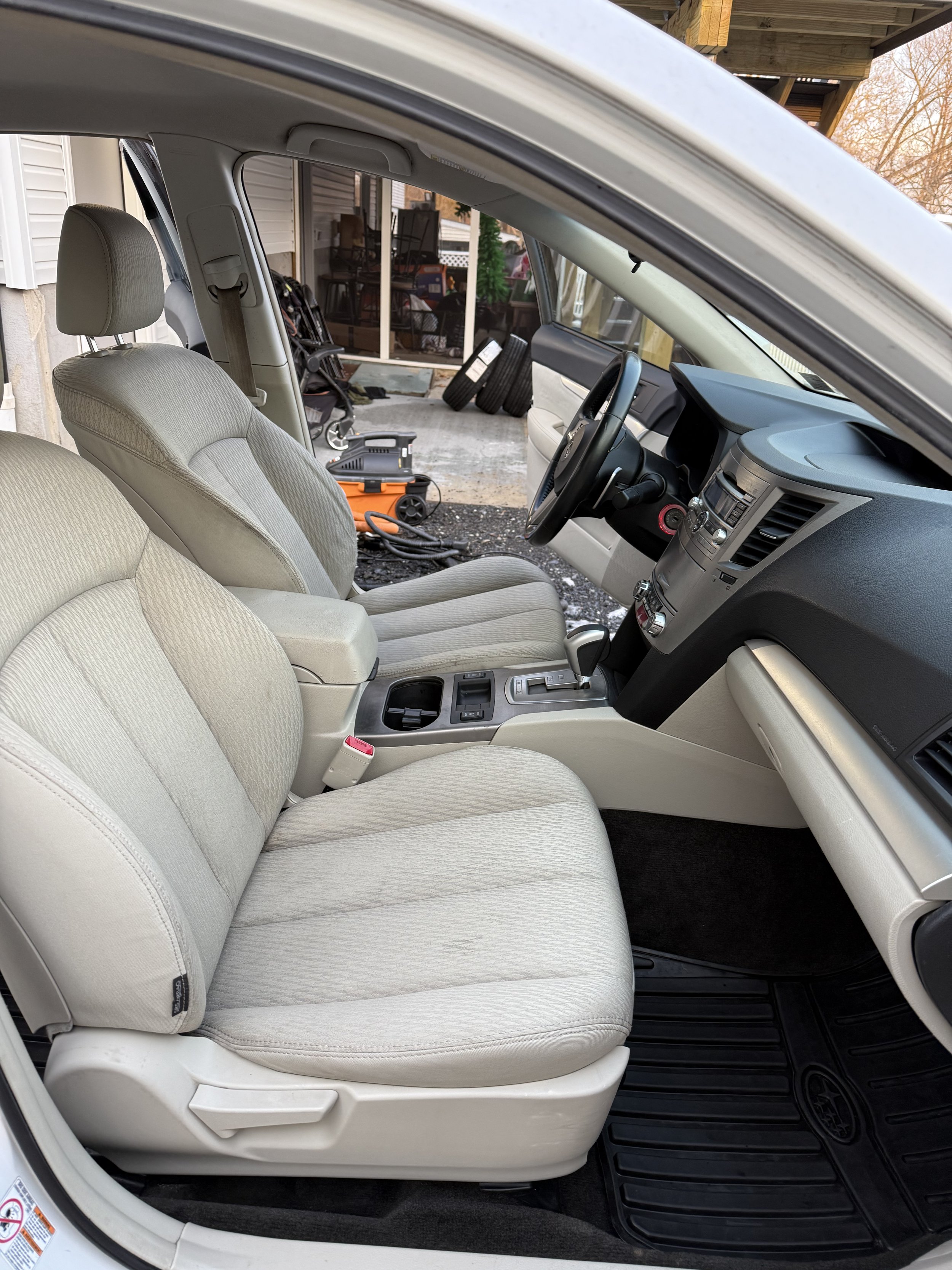 The interior of a beige car showing the front seats, dashboard, and center console, with a view of a garage and some tools outside.