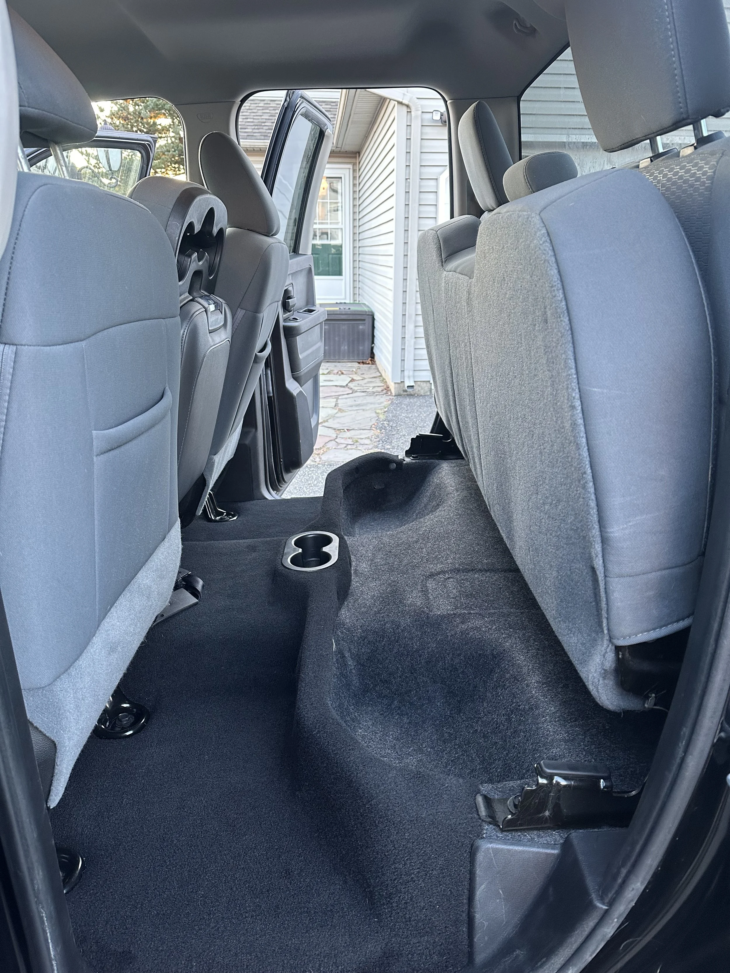 Interior view of a vehicle with the rear seats folded up, showing the spacious cargo area and seatbacks, with a paver walkway and house exterior visible through the open door.
