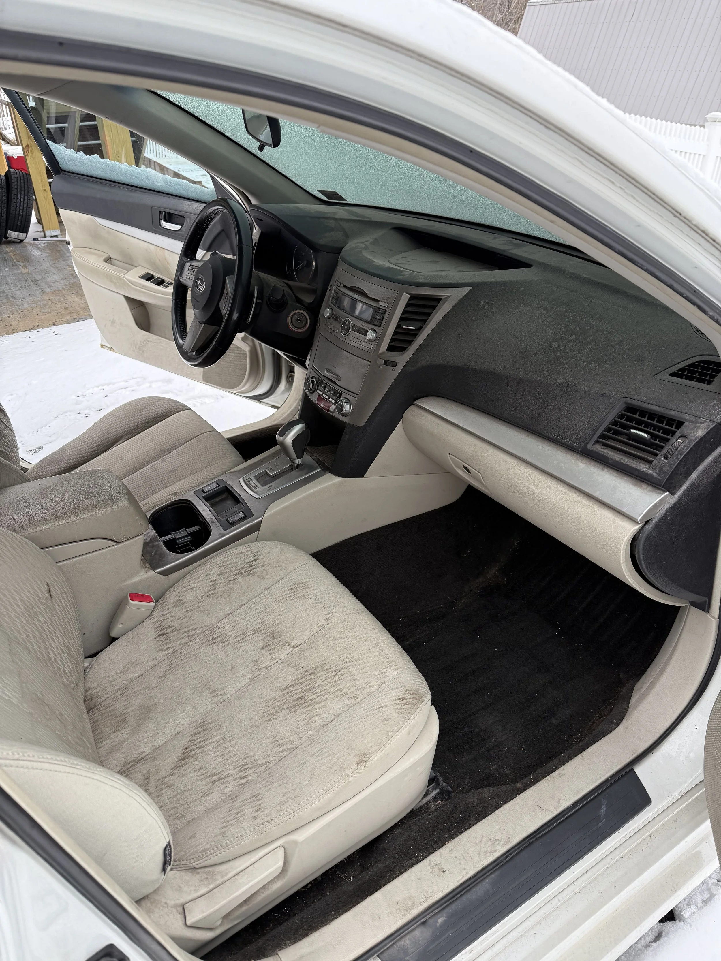 Interior of a white car with a tan fabric seat, black dashboard, and center console, with a view of the snowy ground outside.