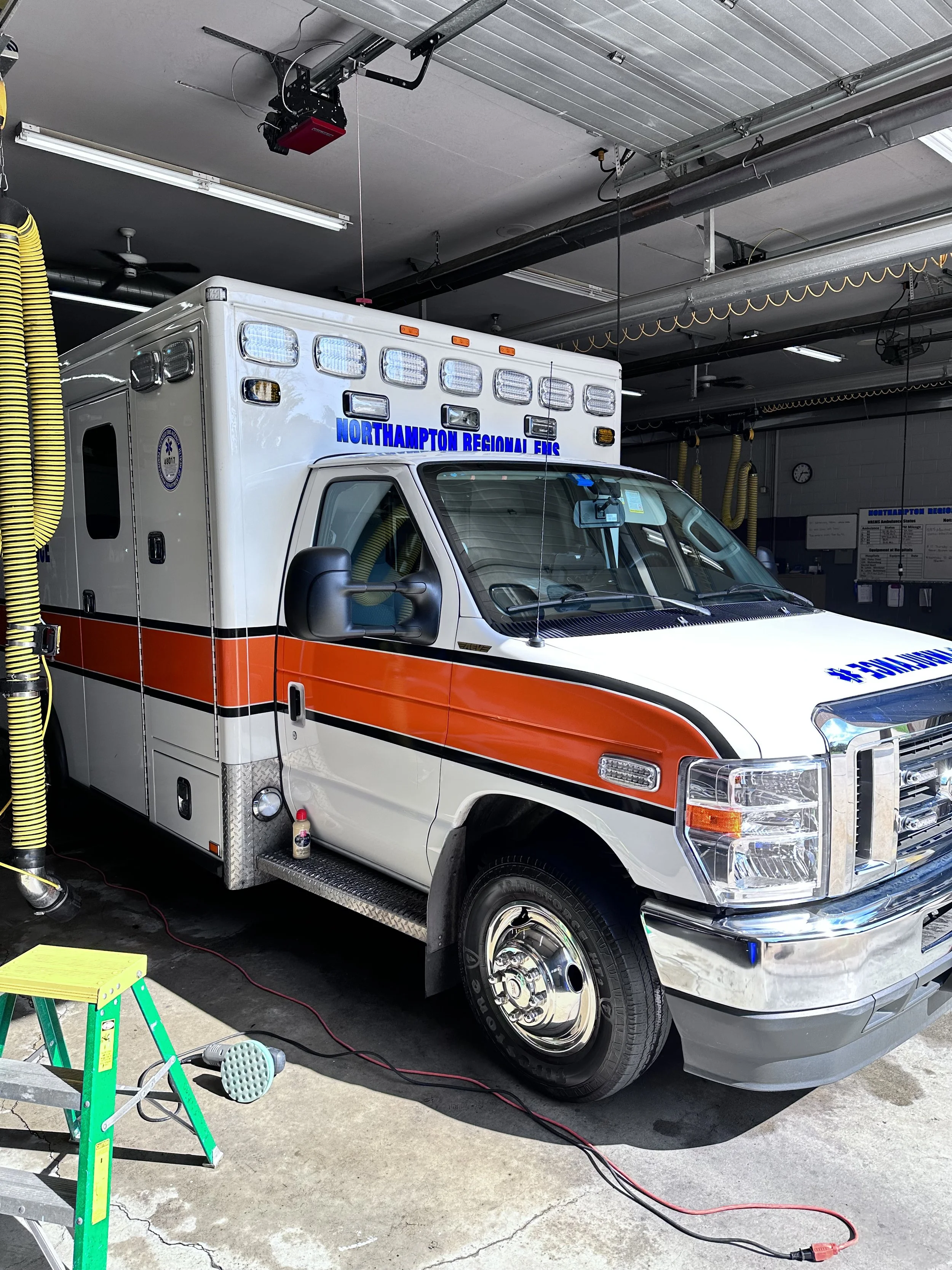 An ambulance from Northampton Regional EMS parked inside a garage with equipment and tools around, including a green step ladder, electrical cords, and a yellow hose.