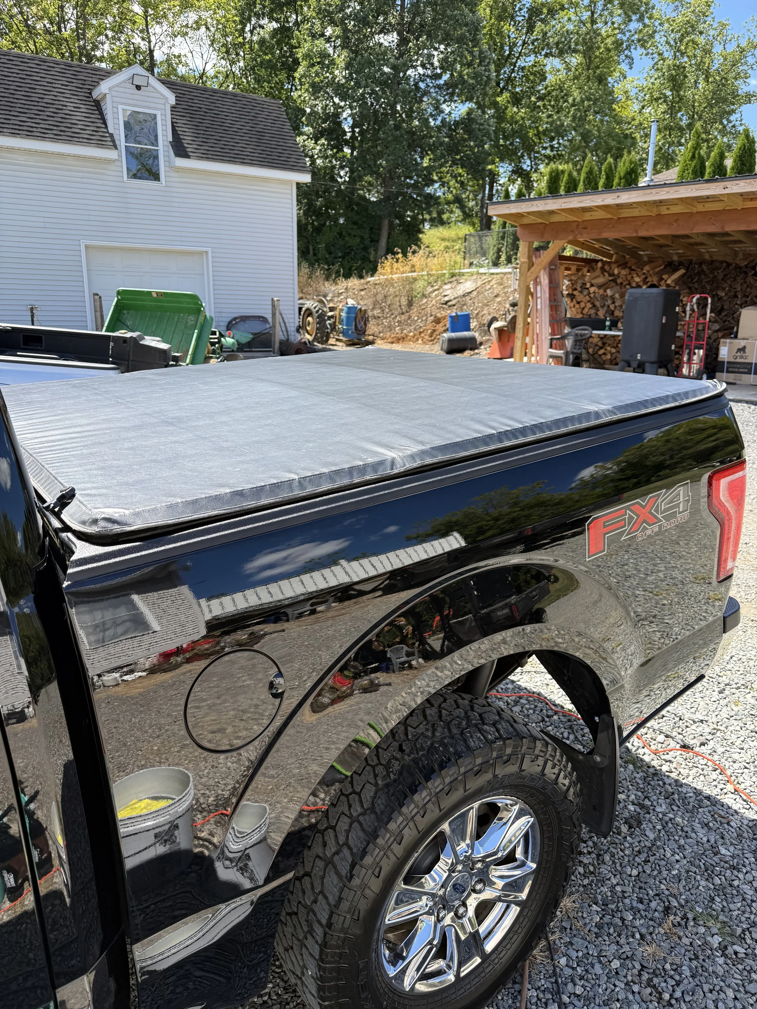 Black pickup truck with a black cover on the bed, parked in a backyard with a white house, woodpile shed, and trees in the background.