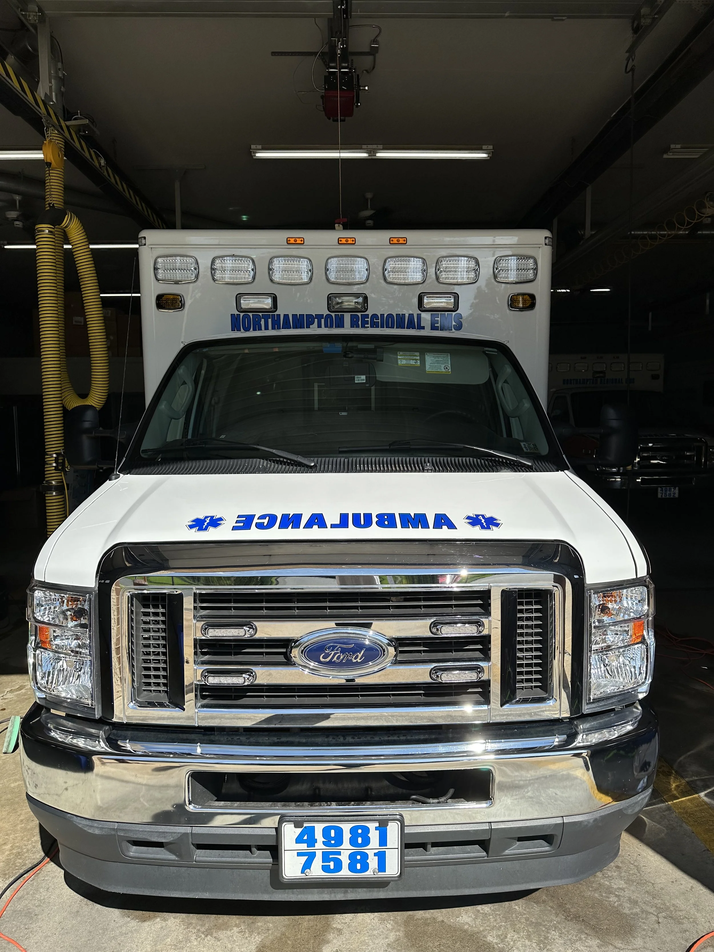 Front view of an ambulance vehicle with 'AMBULANCE' written backward on the hood, a chrome grille with a blue Ford logo, and the words 'NORTHAMPTON REGIONAL EMS' on the top section.