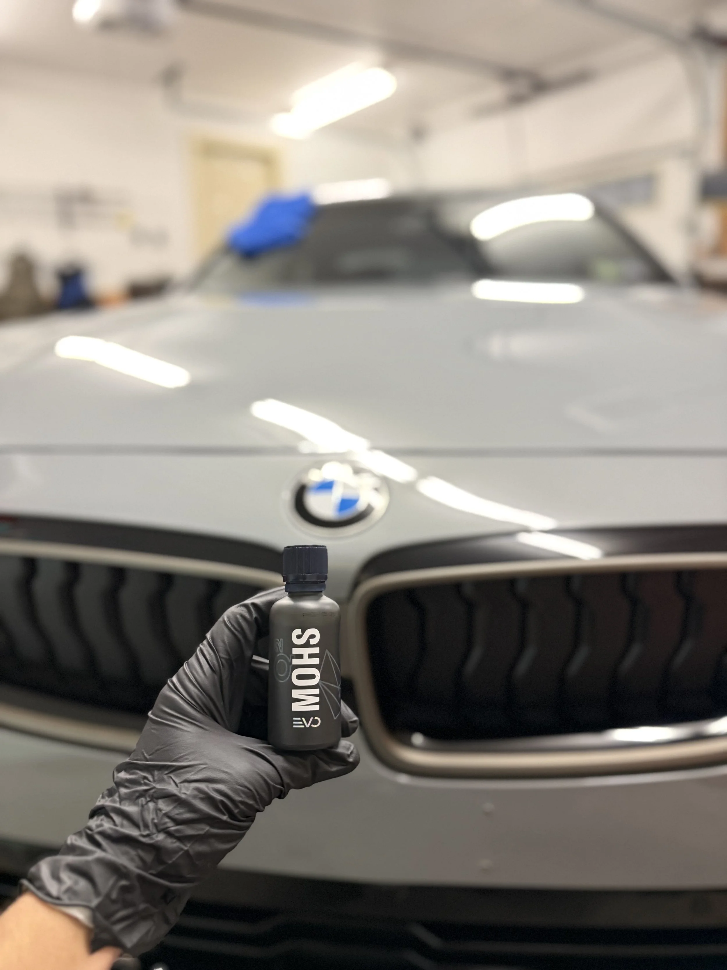 Close-up of a person's hand wearing a black glove holding a small bottle labeled 'SHOWN' in front of a silver BMW car inside a garage or workshop.