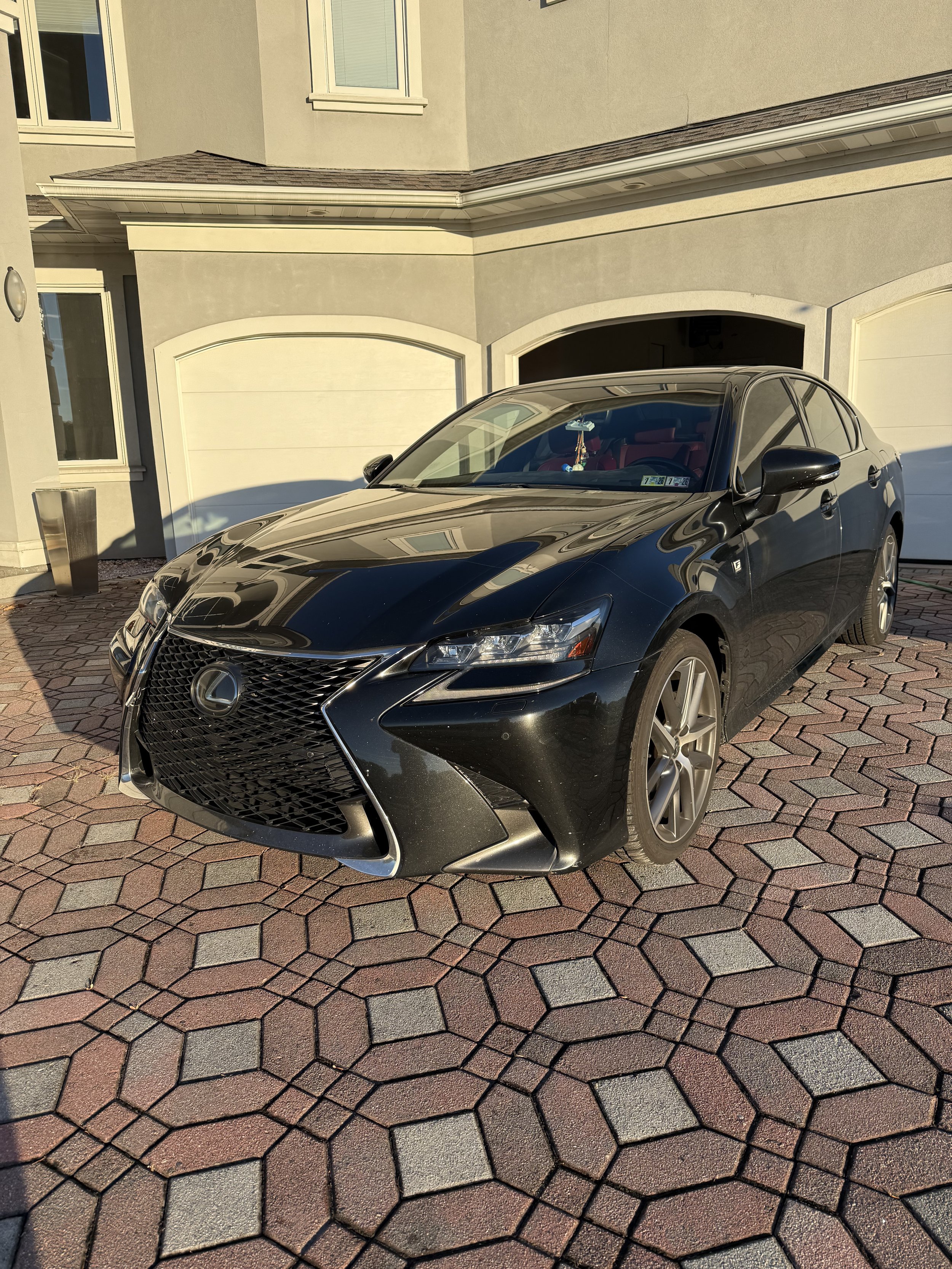 A black Lexus sedan parked on a multi-colored brick driveway in front of a beige house with white garage doors and windows.