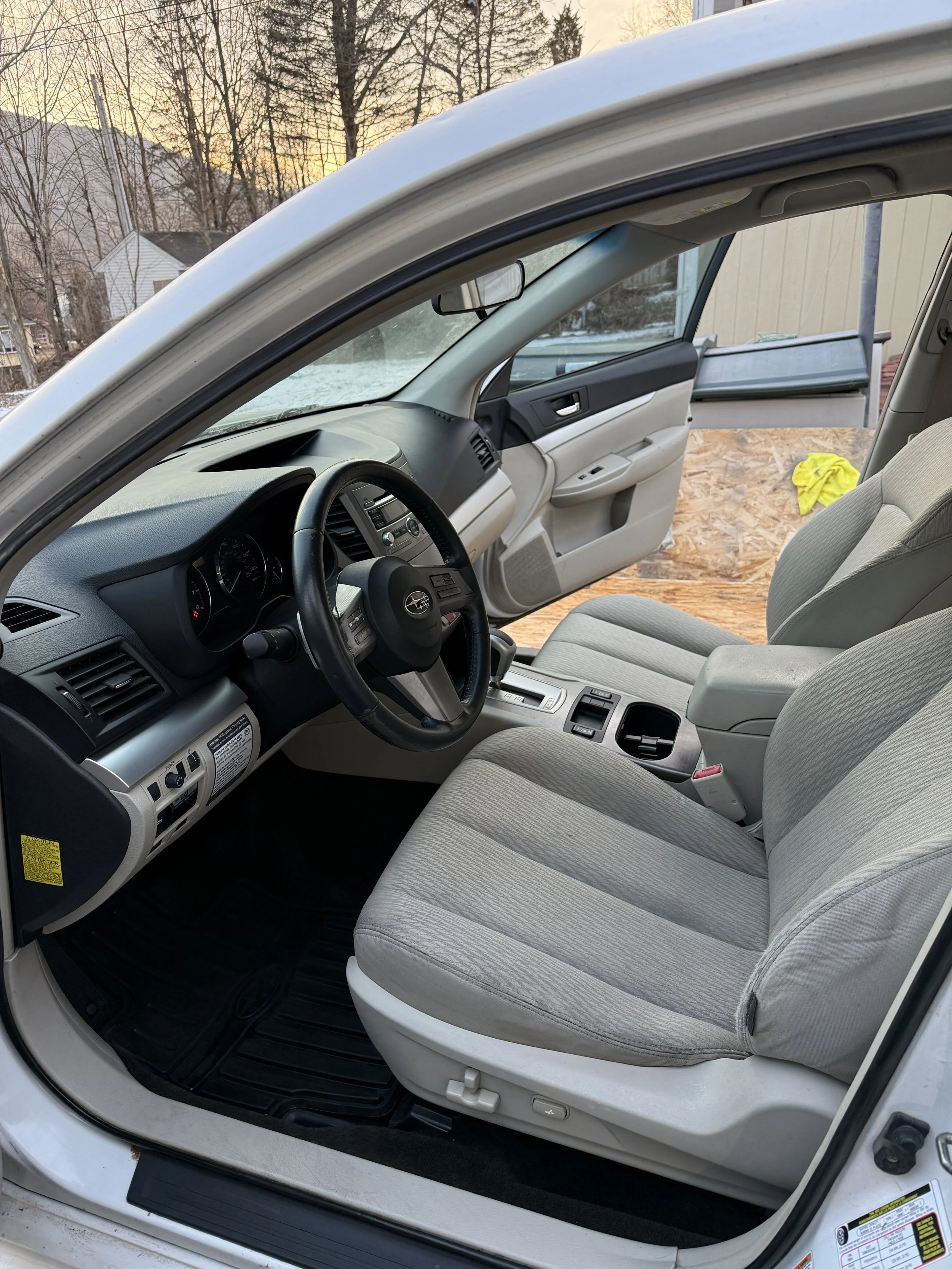 The interior of a silver car parked outdoors during sunset, showing driver's seat, dashboard, steering wheel, and an open passenger door with a view of trees and houses in the background.