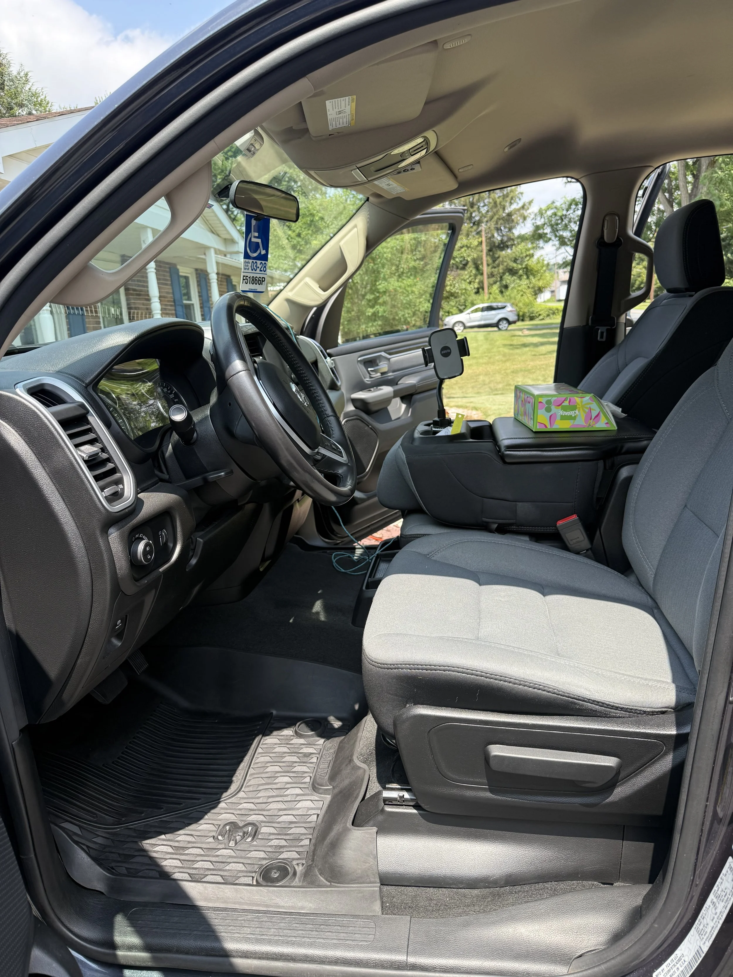 Interior of a pickup truck with a dashboard, steering wheel, front seats, and a center console. There is a box of tissues on the center console. The passenger door is open, revealing a grassy yard and trees outside.