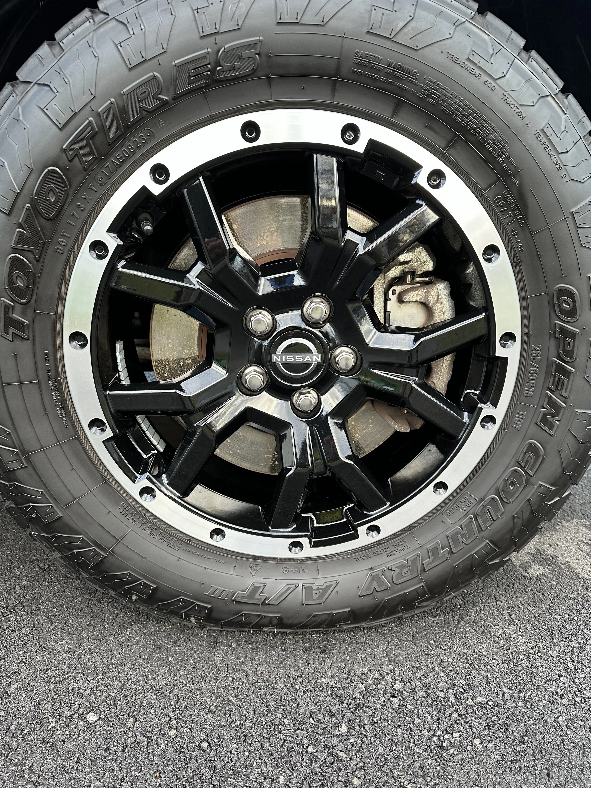 Close-up of a car wheel featuring a black alloy rim with that Nissan logo, mounted with a Toyo Open Country A/T tire.