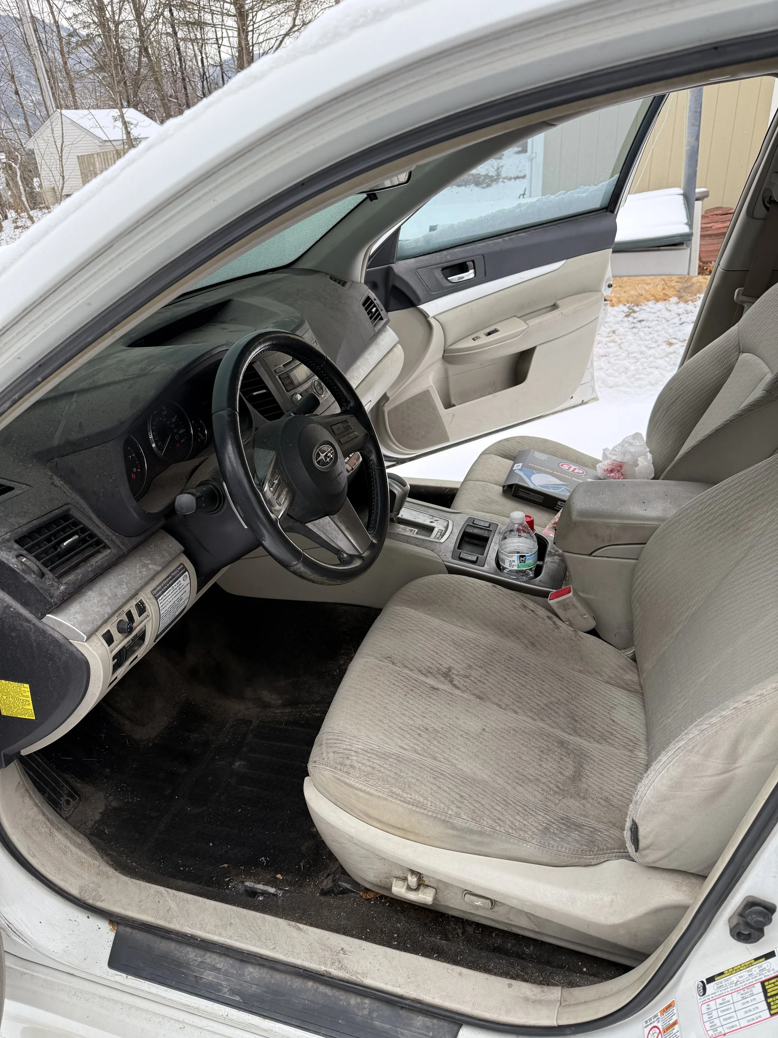 Inside a car with a dirty, beige fabric seat and dashboard, with snow visible outside the open door, containing a water bottle, a pack of tissues, a box, and some snow on the floor.