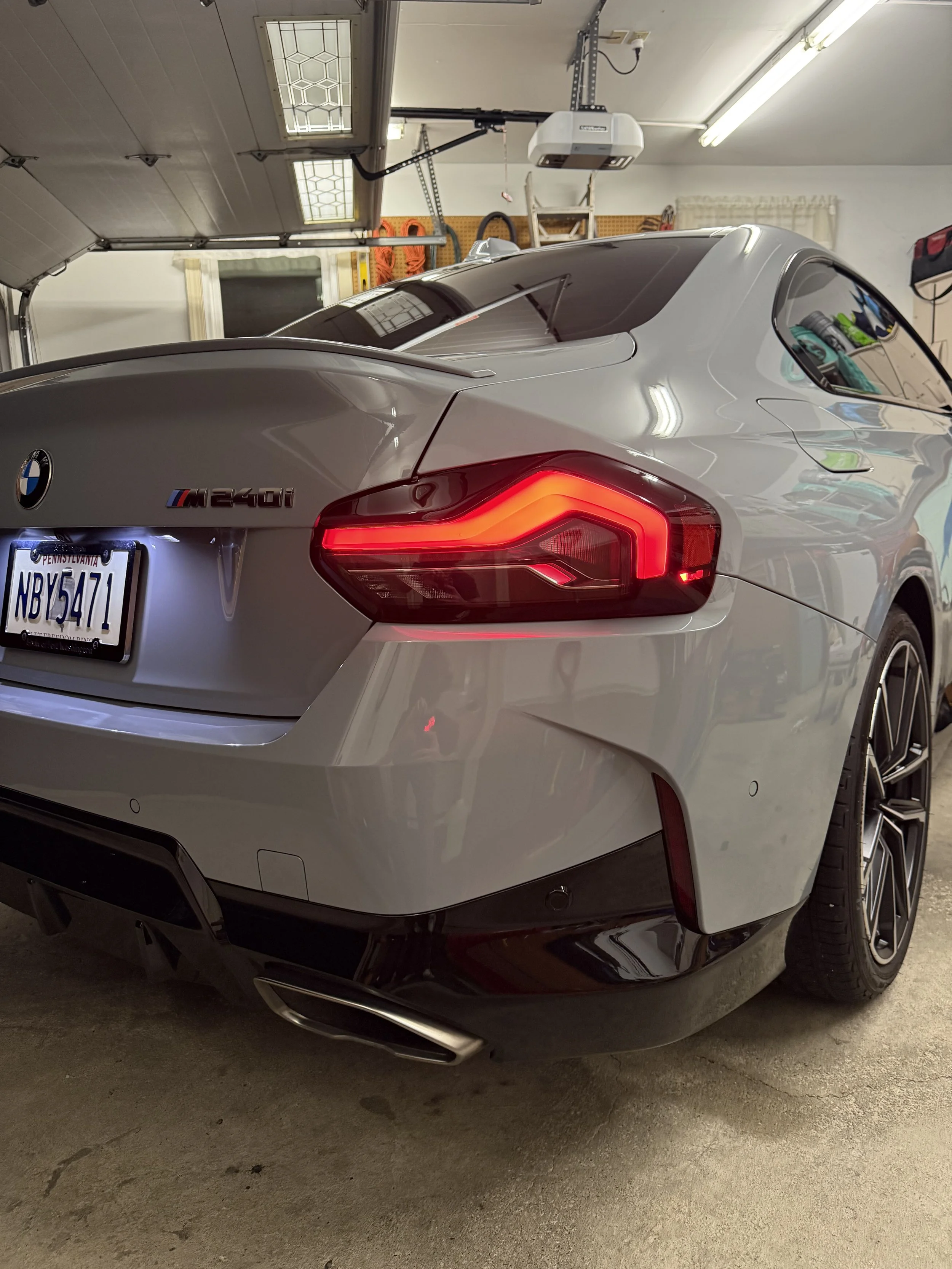 The rear view of a silver BMW M240i parked in a garage, showing the tail lights, license plate, and dual exhaust, with garage tools and equipment visible in the background.