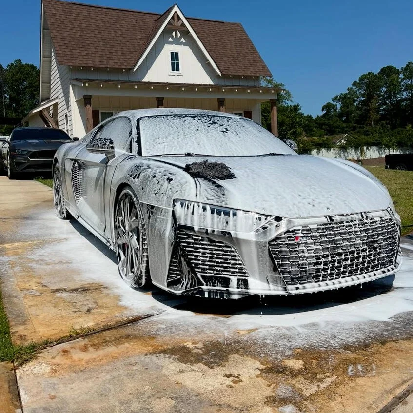 A silver sports car covered in soap suds, parked in front of a house, with foam and water on the driveway.