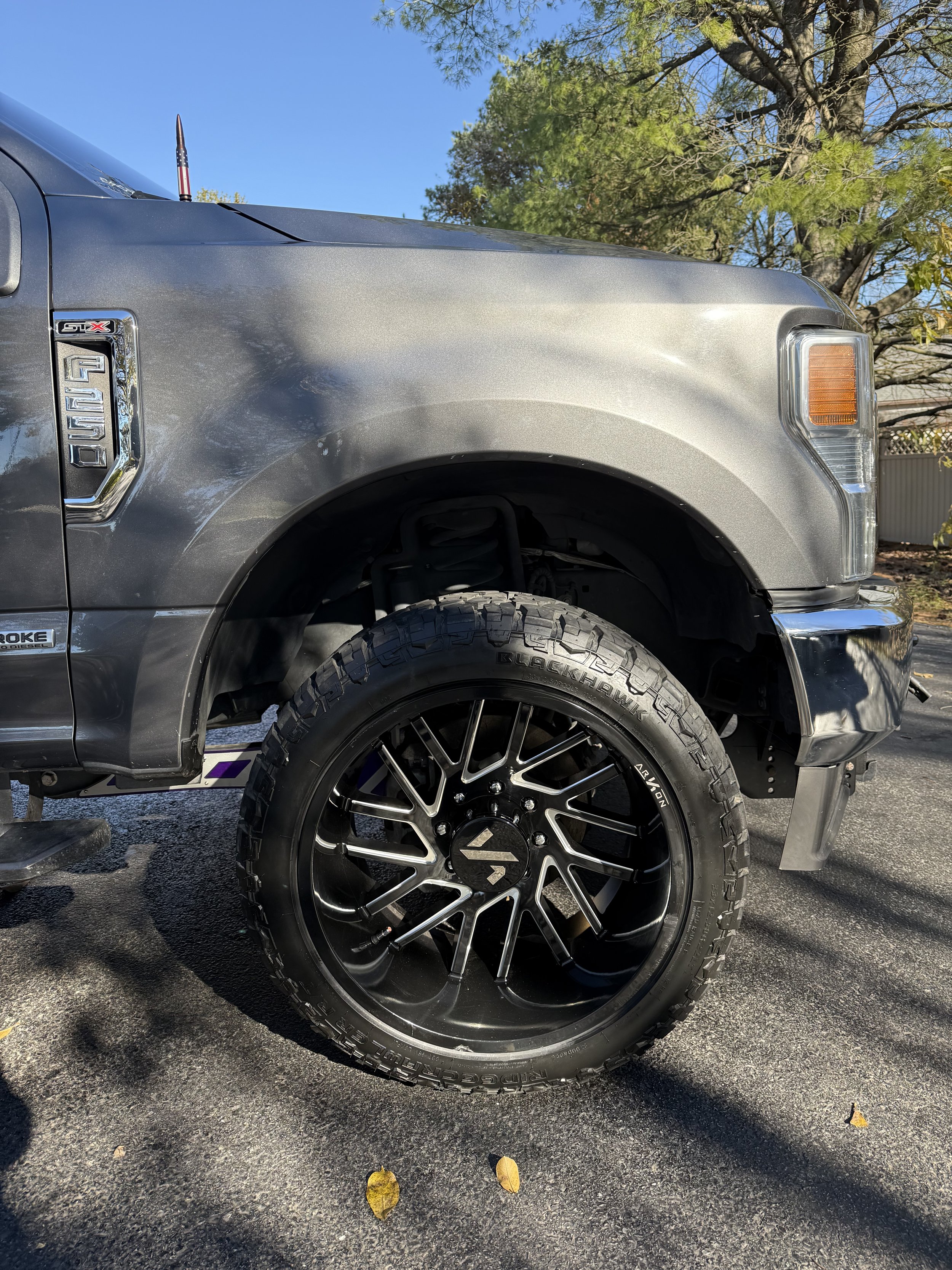 Close-up of the front left side of a gray pickup truck, showing the wheel, tire, and part of the front fender. The truck has black aftermarket rims and off-road tires, parked on a driveway with trees and blue sky in the background.