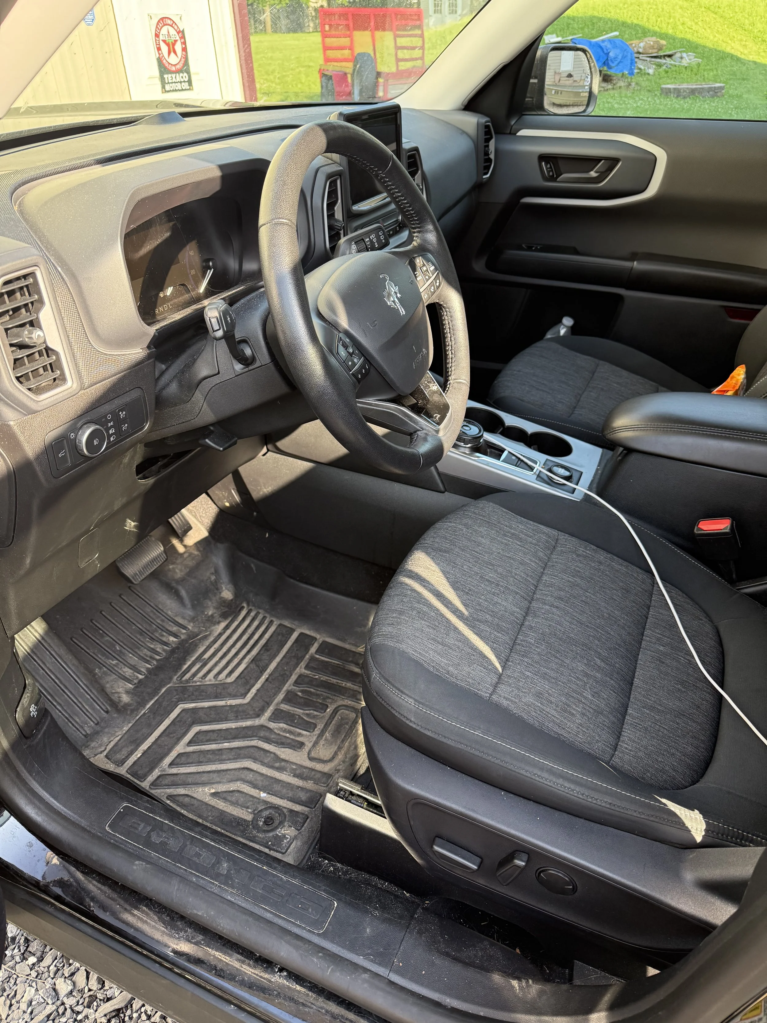 Interior of a vehicle, showing the driver's seat, steering wheel, dashboard, and center console. The floor mat has dirt and debris, and a charging cable is plugged into a port on the side of the seat.