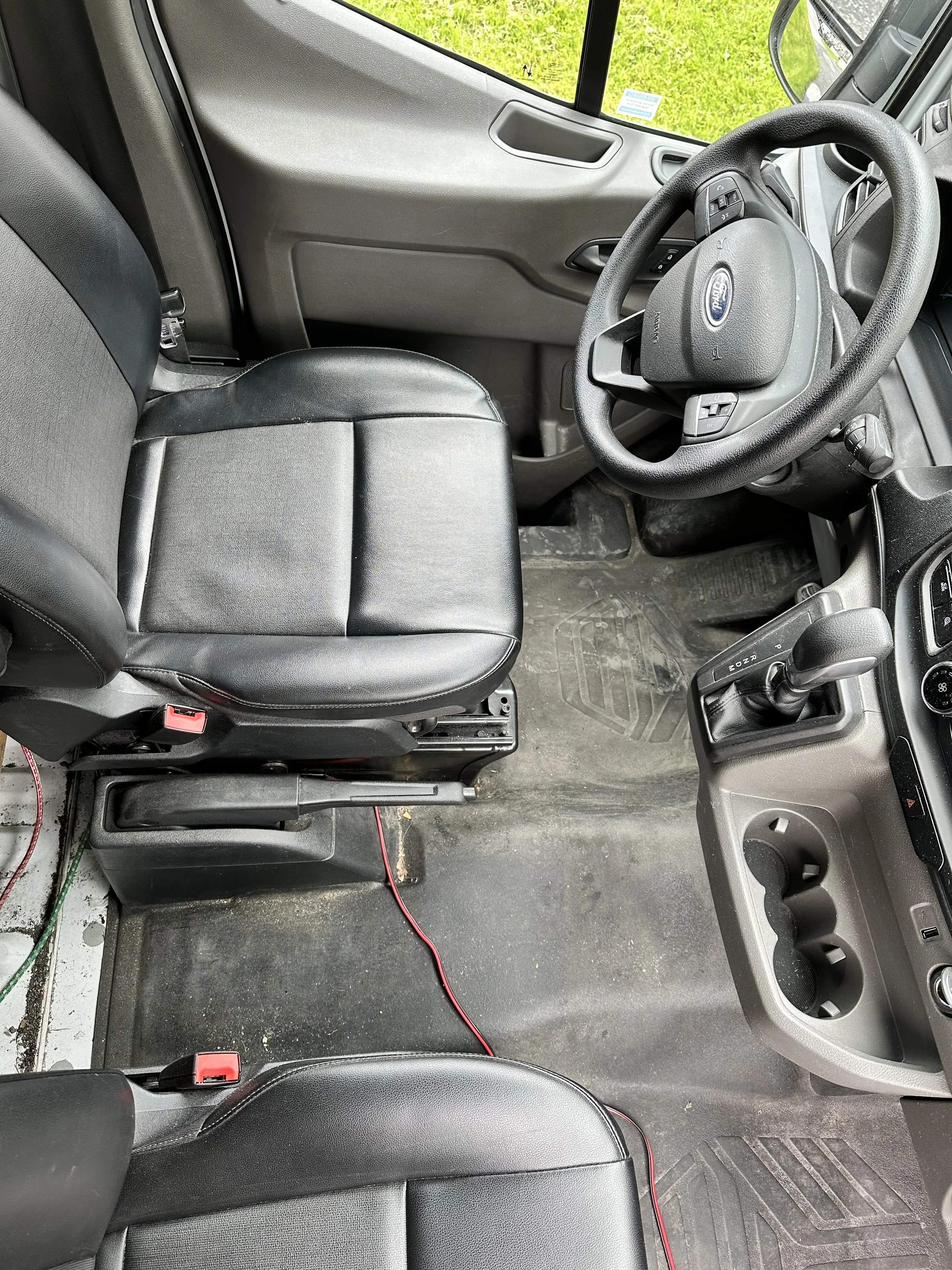 Interior view of a commercial vehicle's driver and passenger seats, steering wheel, gear shift, and dashboard, with rubber floor mats and some wires on the floor.