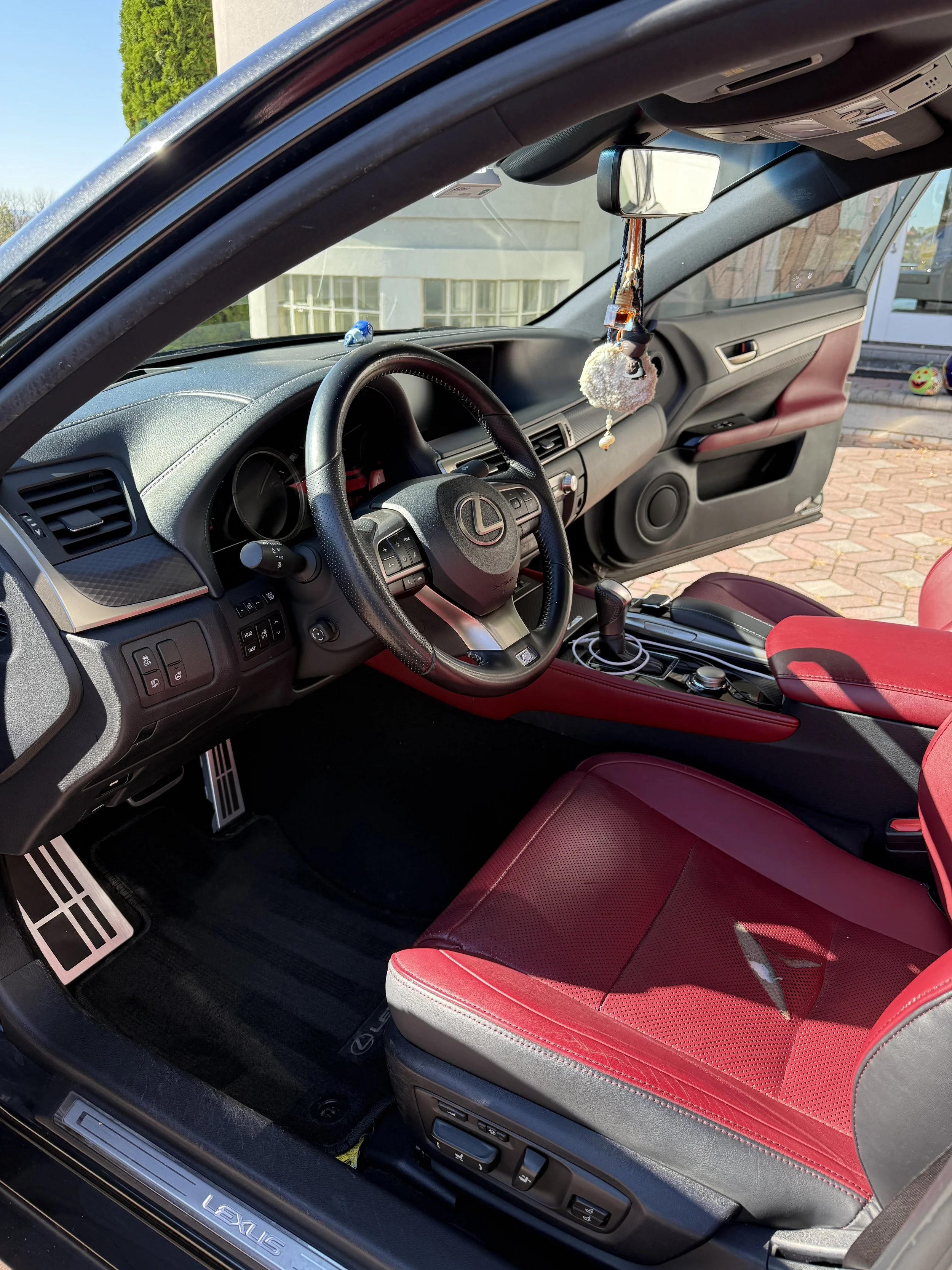 Interior of a Lexus car with a black dashboard, red leather seats, and a steering wheel with the Lexus logo. The rearview mirror has hanging decorations, and the car is parked outside on a sunny day.