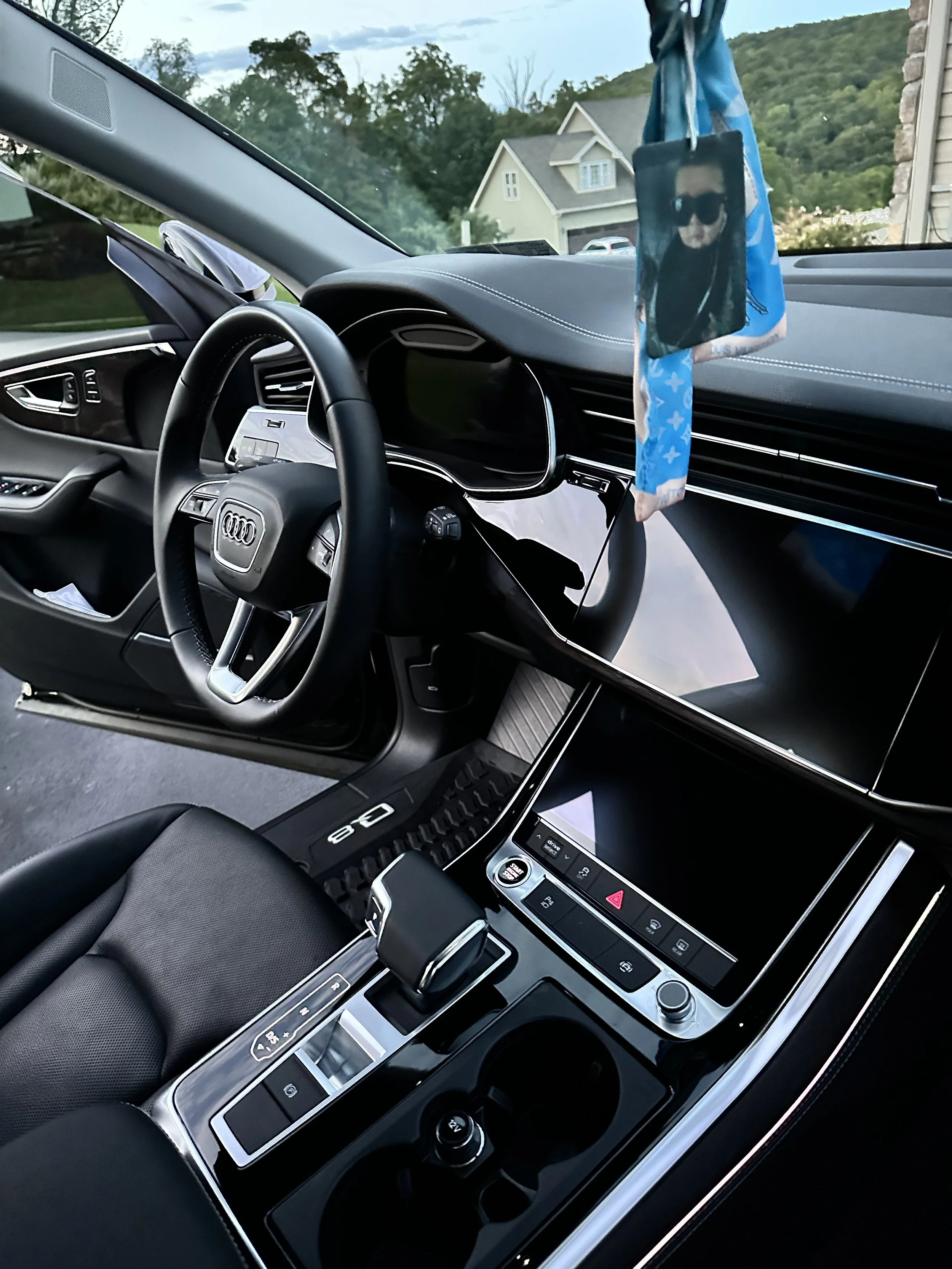 Interior of a modern car, showing the steering wheel, digital dashboard, gear shift, center console with touchscreen, and a hanging air freshener or decoration from the rearview mirror. Houses and trees are visible through the windshield.