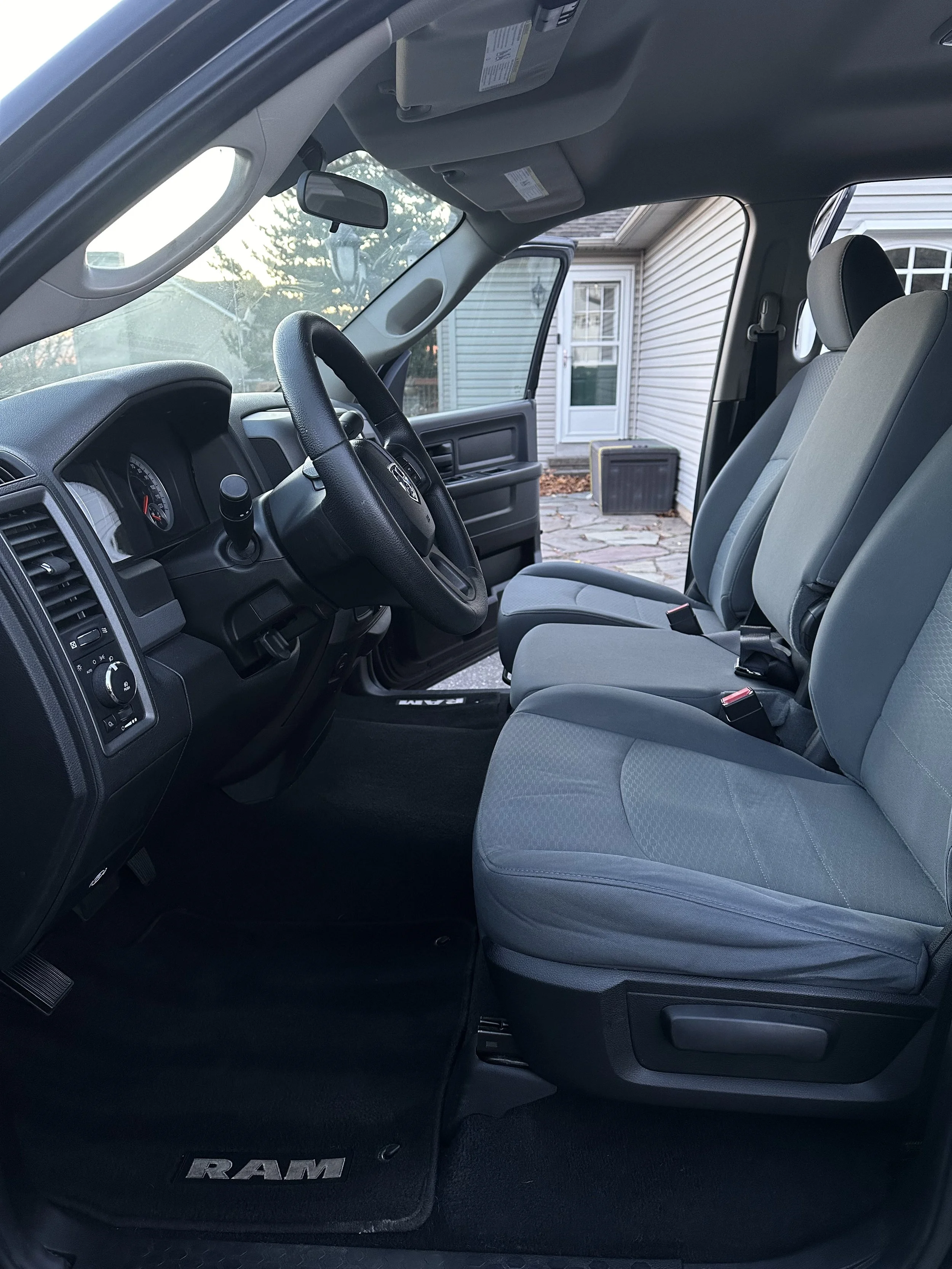 Interior view of a vehicle's front cabin, showing the steering wheel, dashboard, front passenger seat, and floor mat with the RAM logo. The vehicle is parked outside near a house with visible siding and a window.
