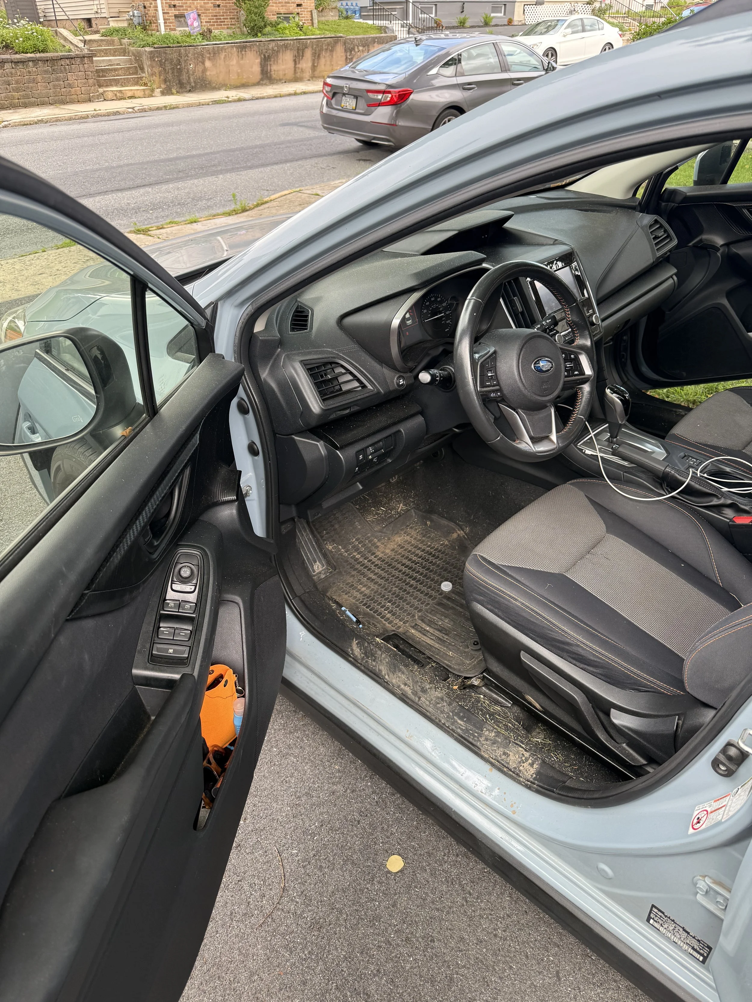 Inside view of a car with the driver's side door open, showing the steering wheel, dashboard, driver's seat with dirt and debris on the floor, and the surrounding outdoor environment with parked cars and a brick house.