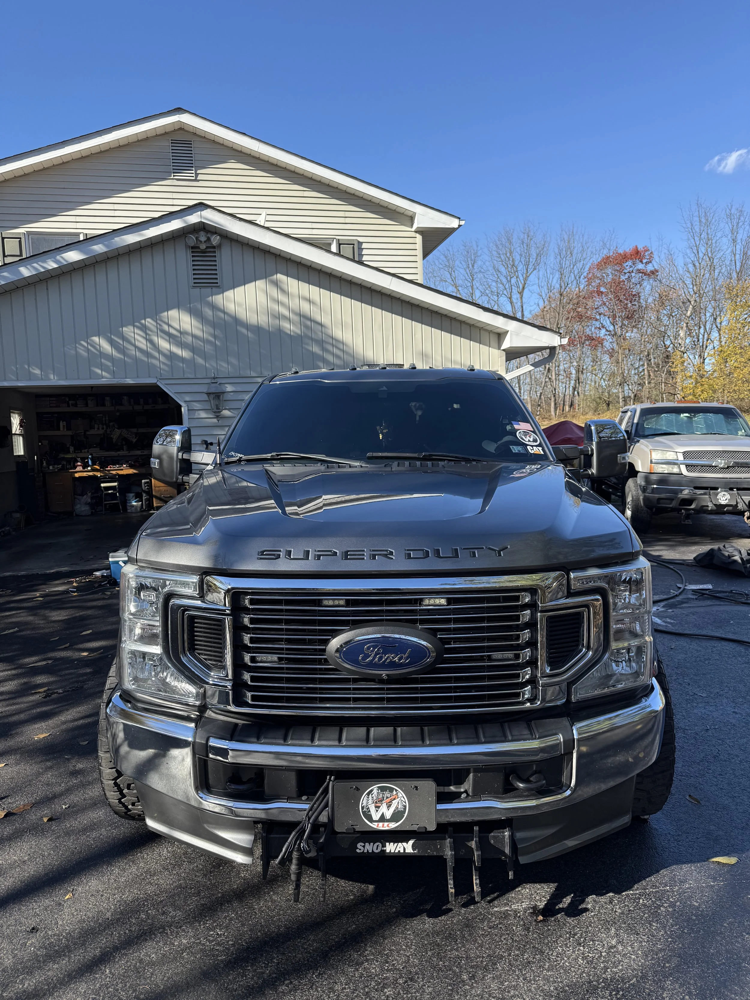 Front view of a black Ford Super Duty pickup truck parked in a driveway, with garage and other vehicles visible in the background, and a house with beige siding behind it.