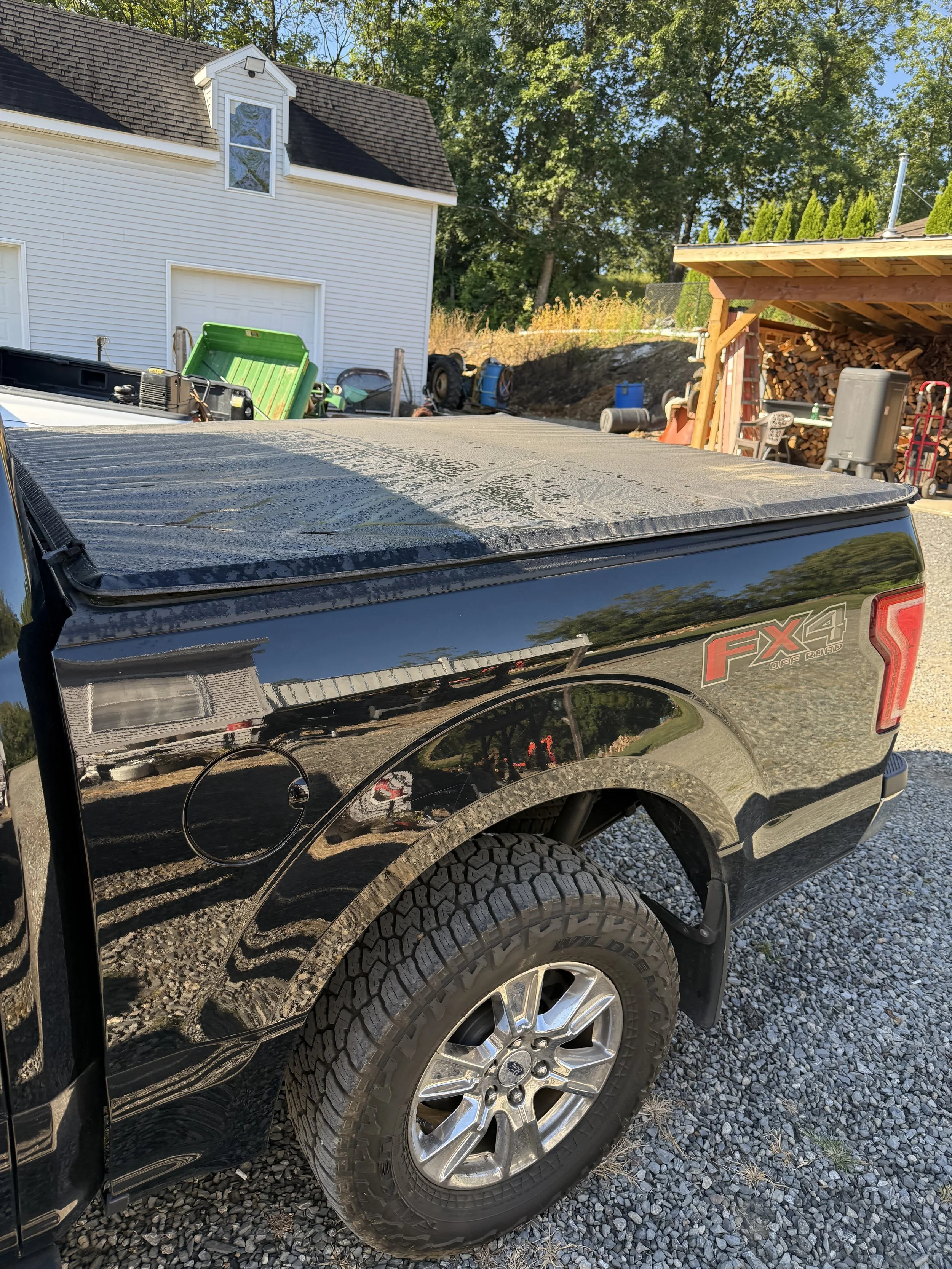 Black pickup truck with an FX4 Off Road decal parked on a gravel driveway, with a covered bed and a large tire. In the background, there are a white building, some yard tools, and a wood storage shed.