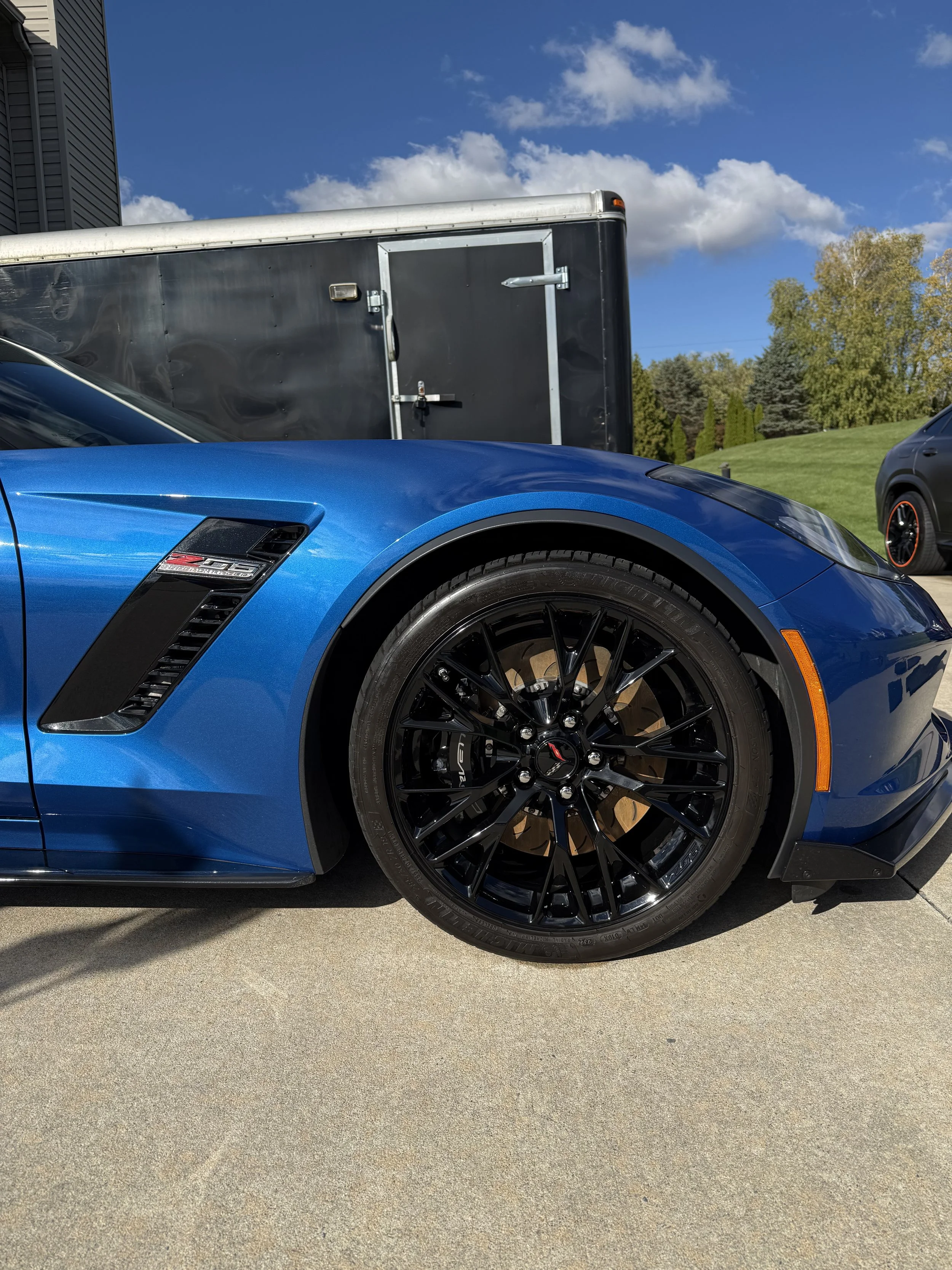 Blue sports car with black wheels parked on a concrete driveway with a black trailer in the background and clear blue sky with a few clouds.