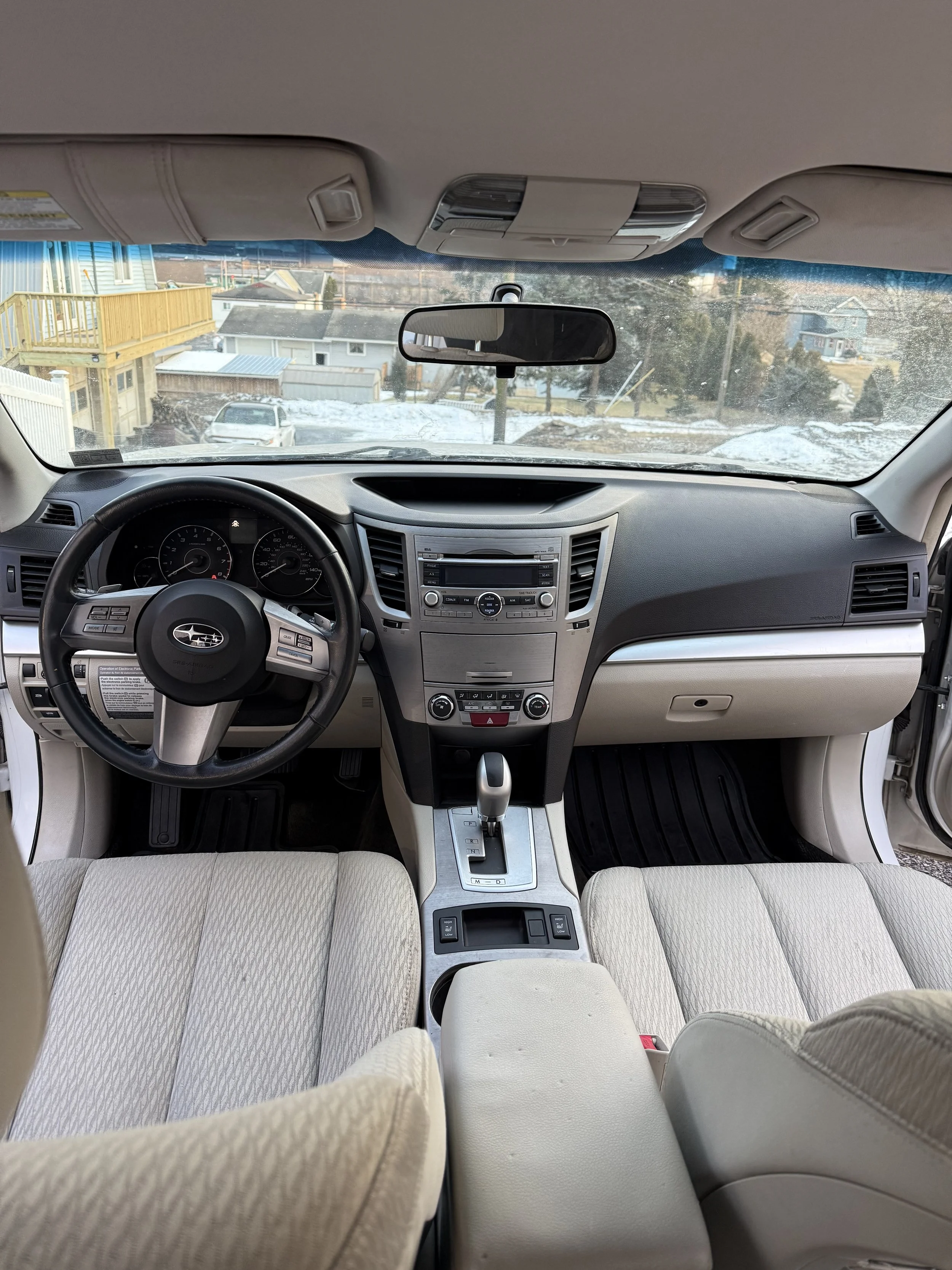 View of a car interior from the back seat showing the dashboard, steering wheel, center console, and front seats inside a vehicle parked in a snowy neighborhood.