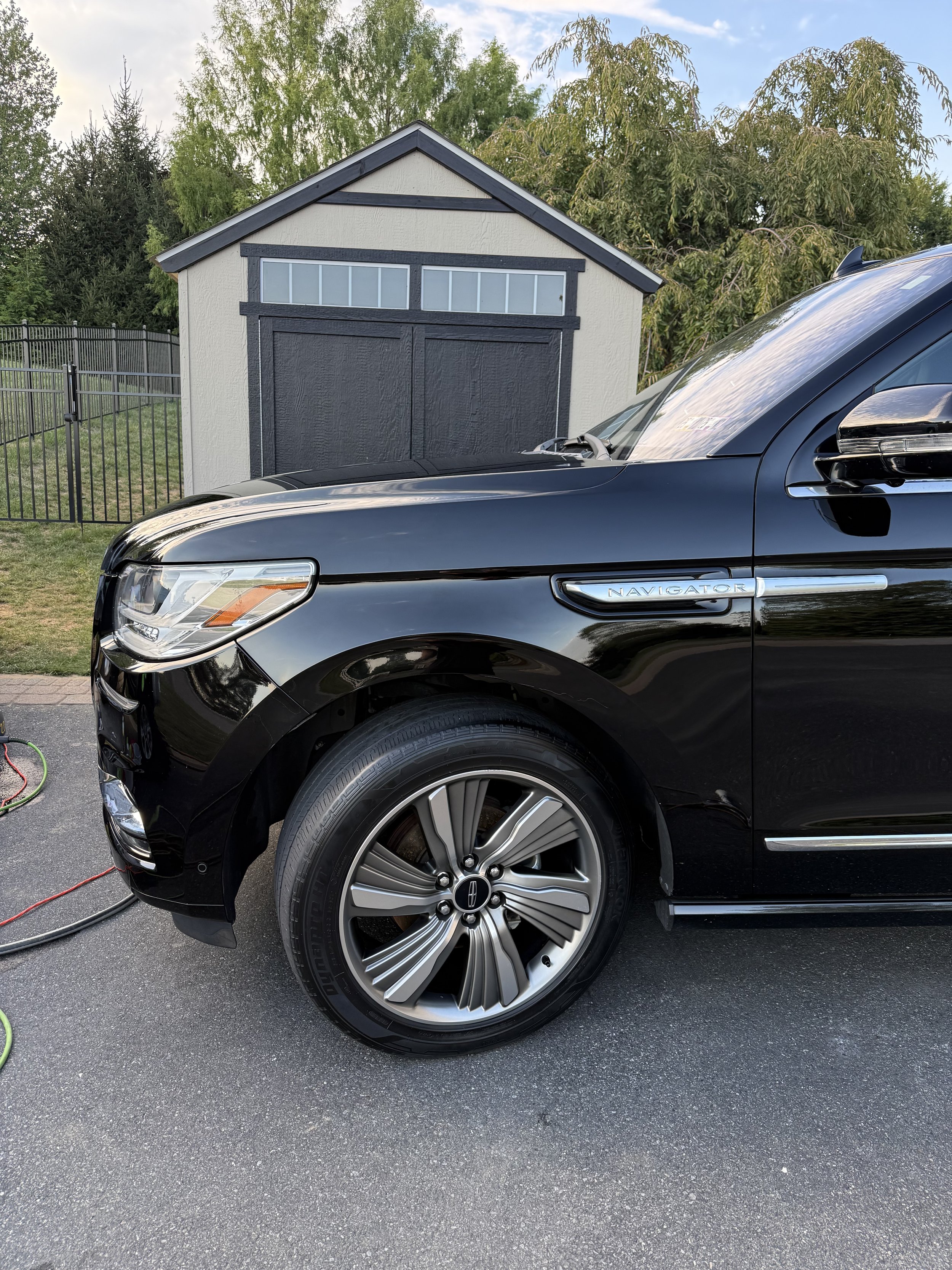 Close-up of the front side of a black Lincoln Navigator SUV parked on a driveway, with a garden shed and trees in the background.