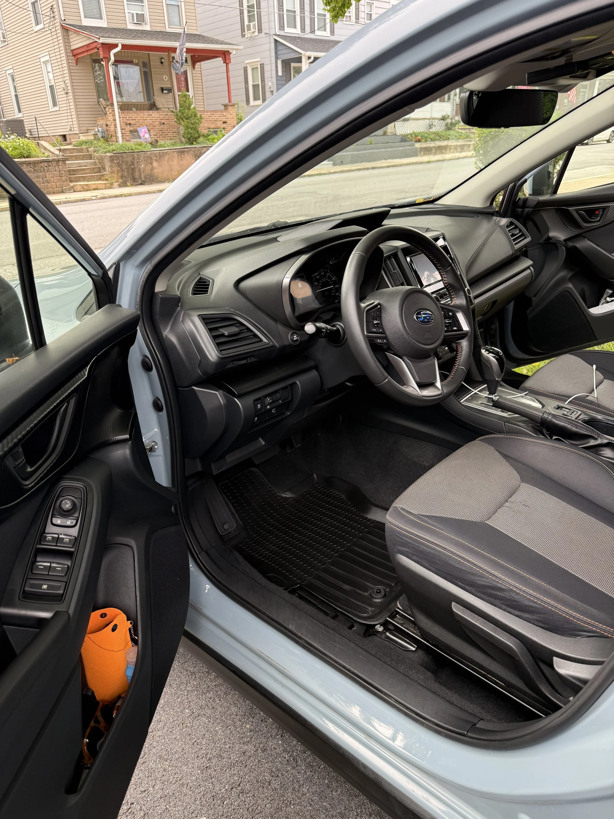 Interior of a modern vehicle, showing the driver's seat, steering wheel, dashboard, and center console, with a residential street view outside.