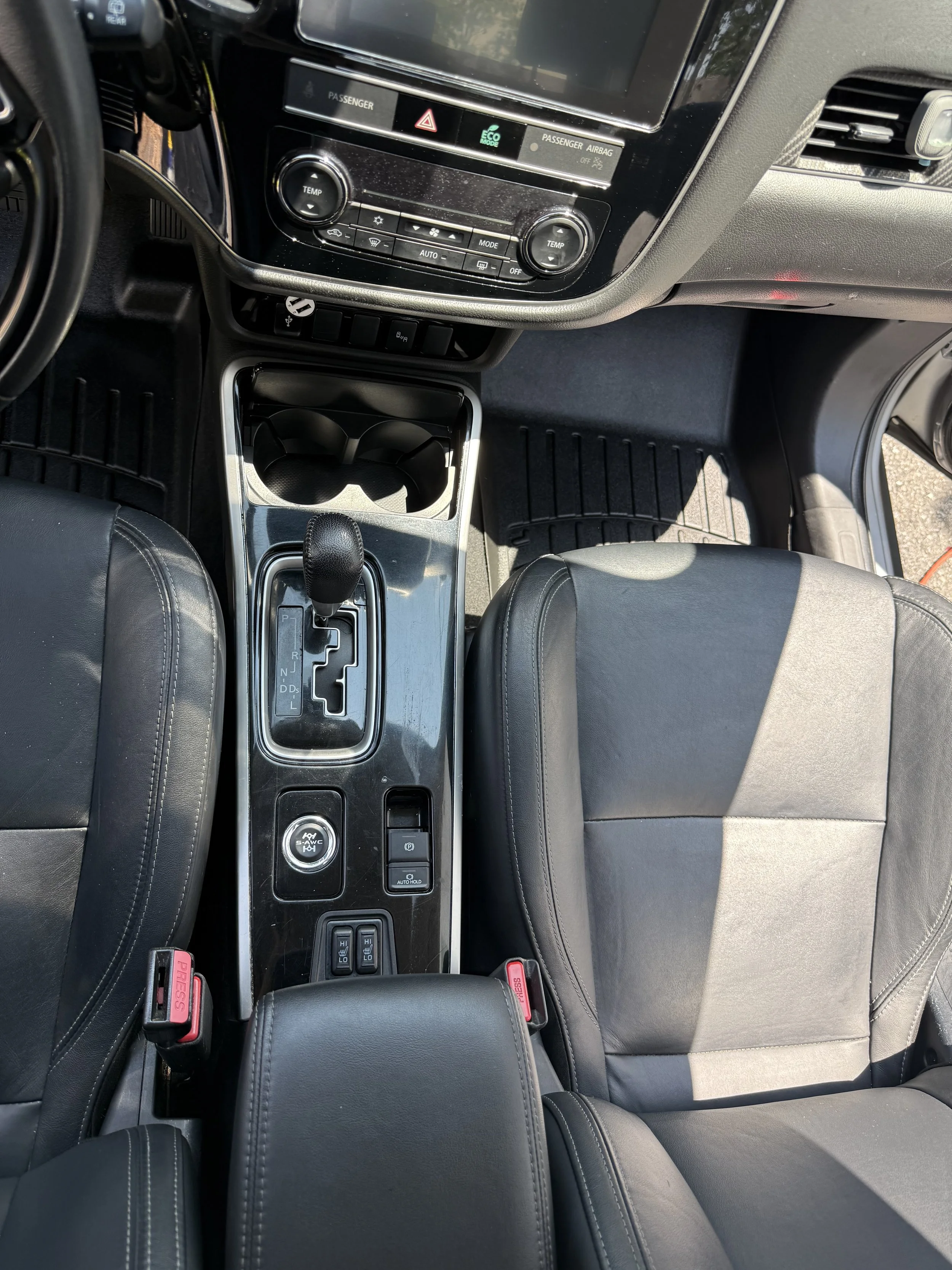 The interior of a vehicle showing the center console with gear shift, cup holders, and climate controls, with black leather seats and a black dashboard.