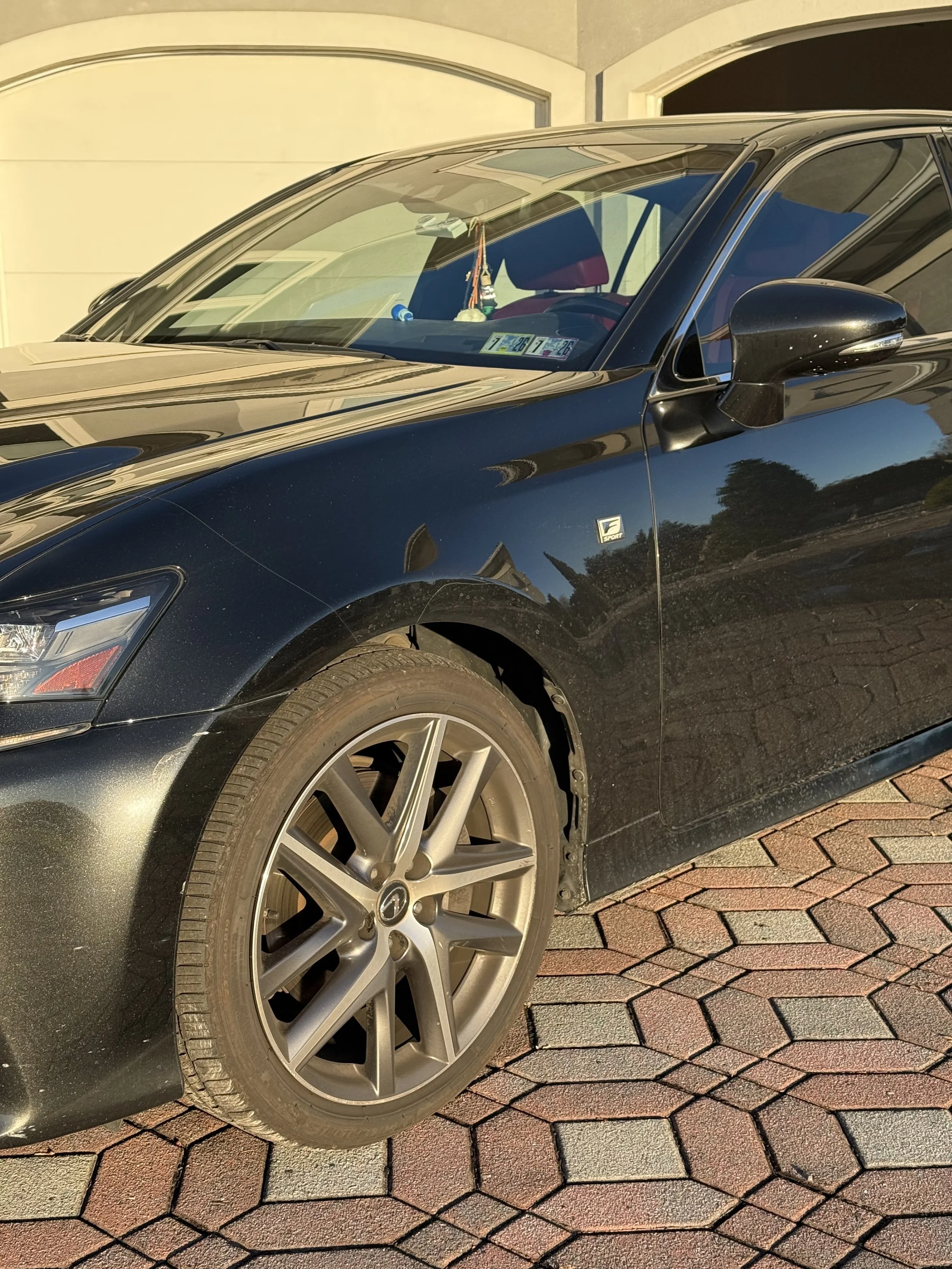 A black Mazda sedan parked on a brick driveway in front of a house, with the front left wheel and part of the side visible, showing some dirt and dust on the car.