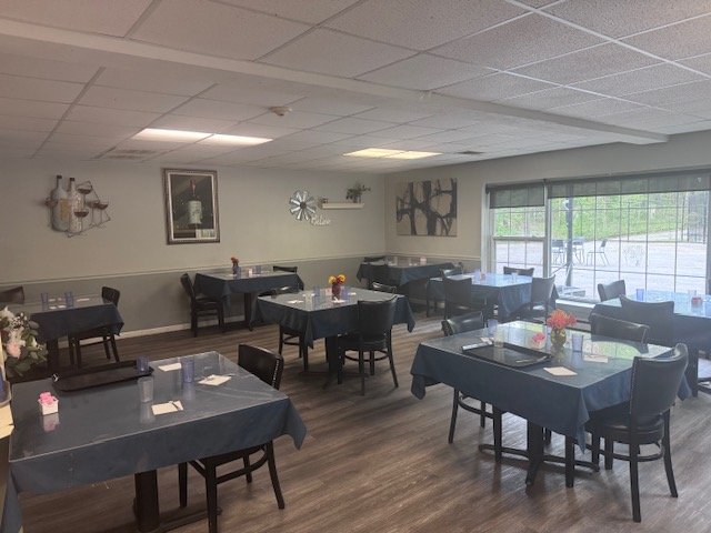 Dining room with several tables covered in dark tablecloths, decorated with small floral centerpieces, set with napkins, glasses, and utensils, and surrounded by black chairs. Large window on the right side provides natural light. Walls are decorated with artwork and a mirror.