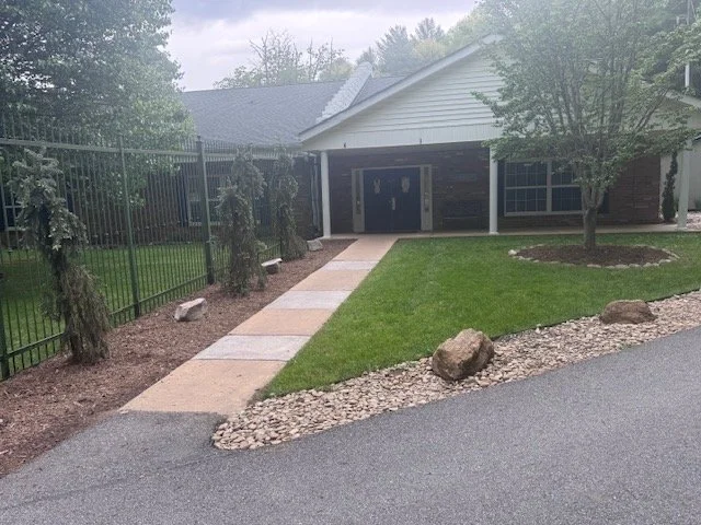 A paved walkway leading to the front door of a house, with a grassy yard, trees, rocks, and a black metal fence.