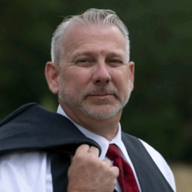 A middle-aged man with short gray hair and a beard, dressed in a white shirt, red tie, and dark suit jacket, holding his suit jacket over his shoulder, outdoors with blurred green foliage in the background.