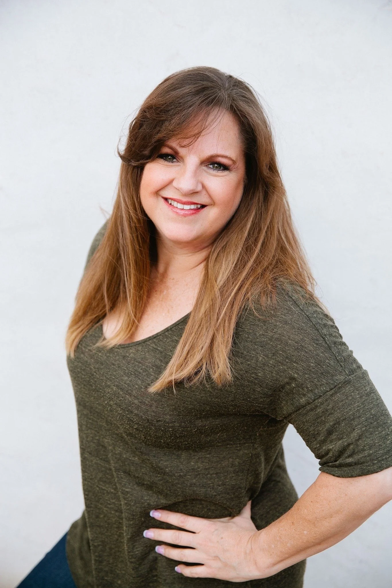 A woman with long, light brown hair and blue eyes, smiling, wearing a green top, standing against a light background with one hand on her hip.