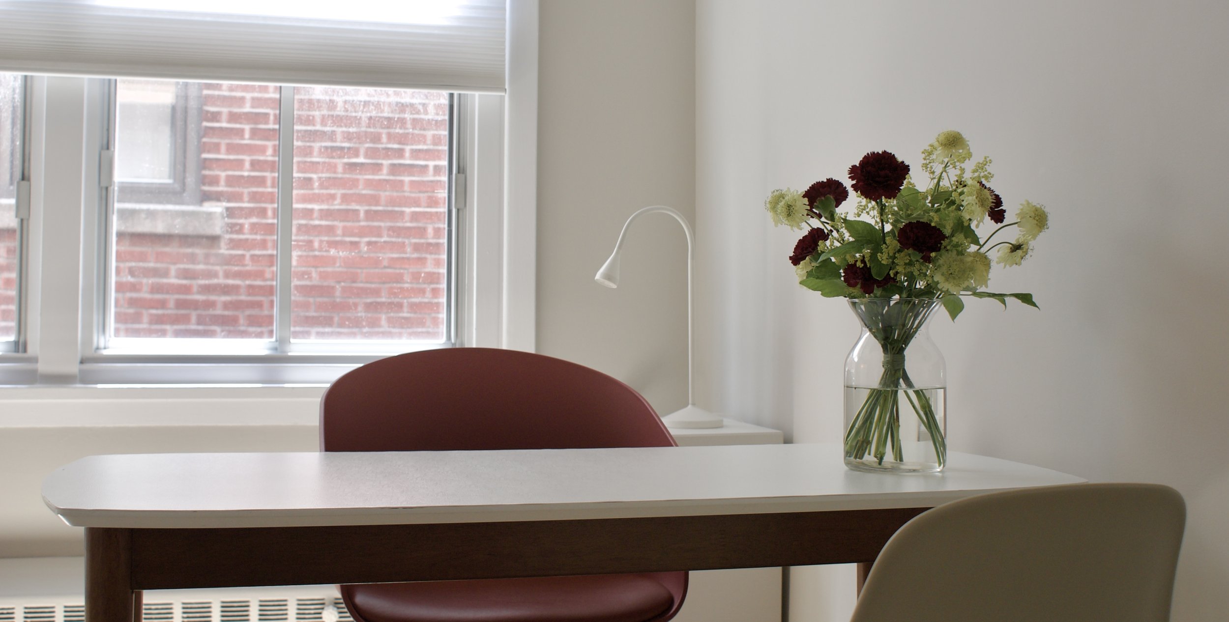 Une fenêtre laissant entrer de la lumière naturelle avec un rideau blanc partiellement baissé, une table blanche avec un vase contenant des fleurs, et une chaise rouge dans une pièce lumineuse et minimaliste.