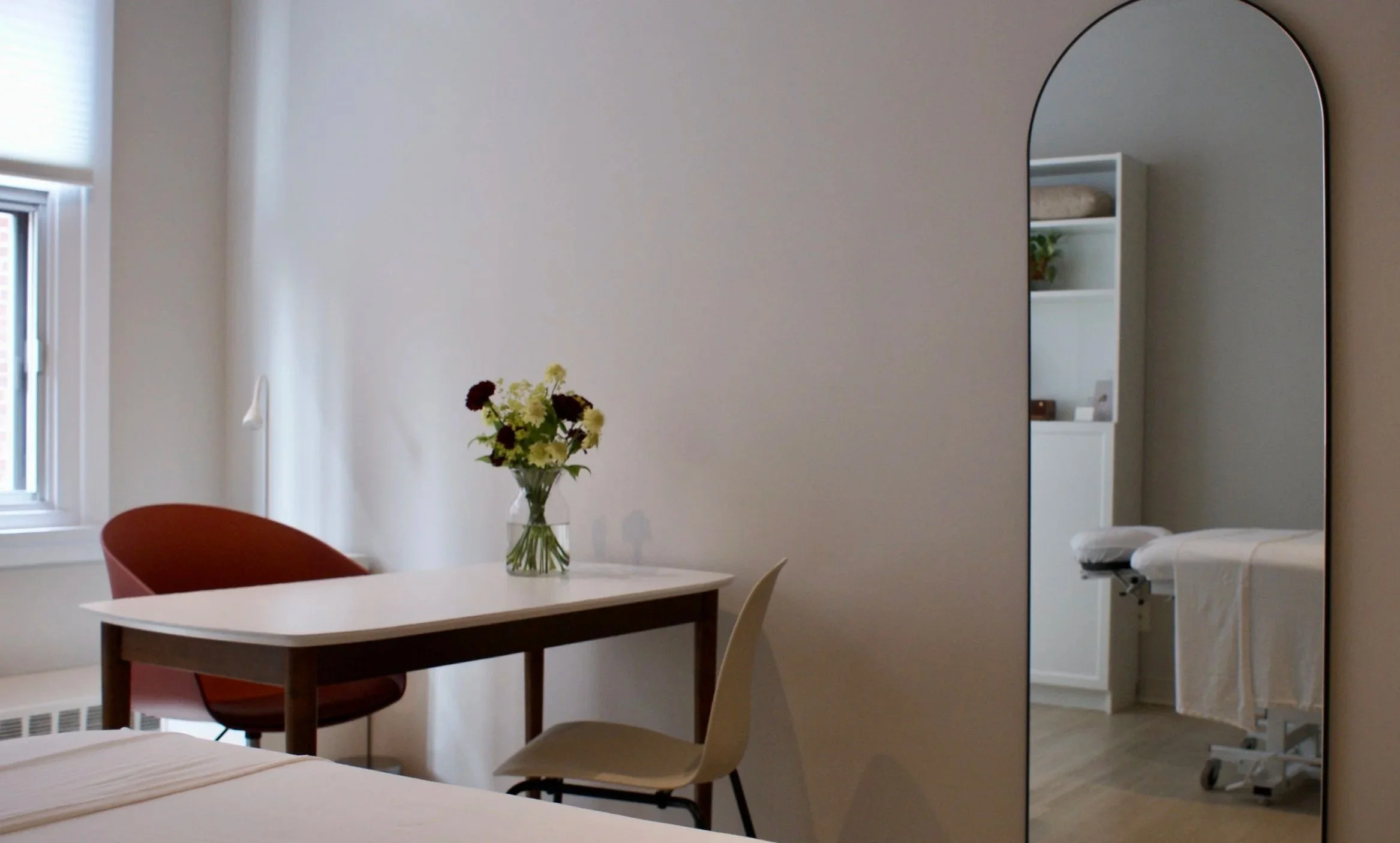 Une pièce intérieure avec une table blanche, une chaise rouge, un vase avec des fleurs blanches et foncées, et un miroir à arqué sur le mur. On voit aussi un lit en partie et une étagère avec des objets.