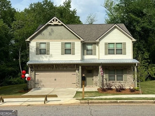 A two-story house with a beige garage door, cream-colored siding, and green window shutters. There is an American flag hanging near the front door and decorative balloons on the left side of the house. The house has a pitched roof and is surrounded b