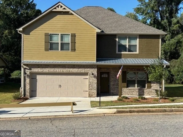 A two-story house with a yellowish-brown upper section, brick lower section, gray garage door, front porch with a small American flag, and a small front yard with a tree.