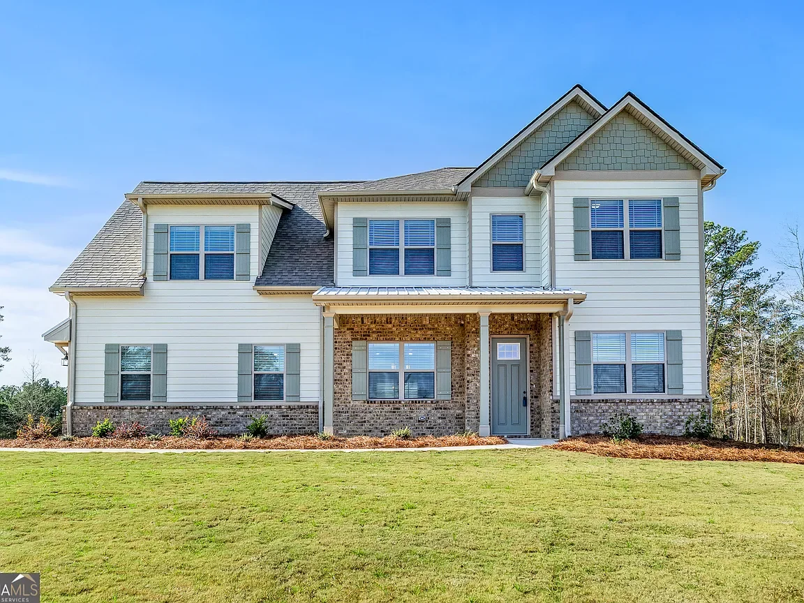 A large two-story suburban house with beige siding, gray shutters, a brick foundation, and a front porch with two columns. The house has multiple windows and a gray front door. The lawn is green and the sky is clear.
