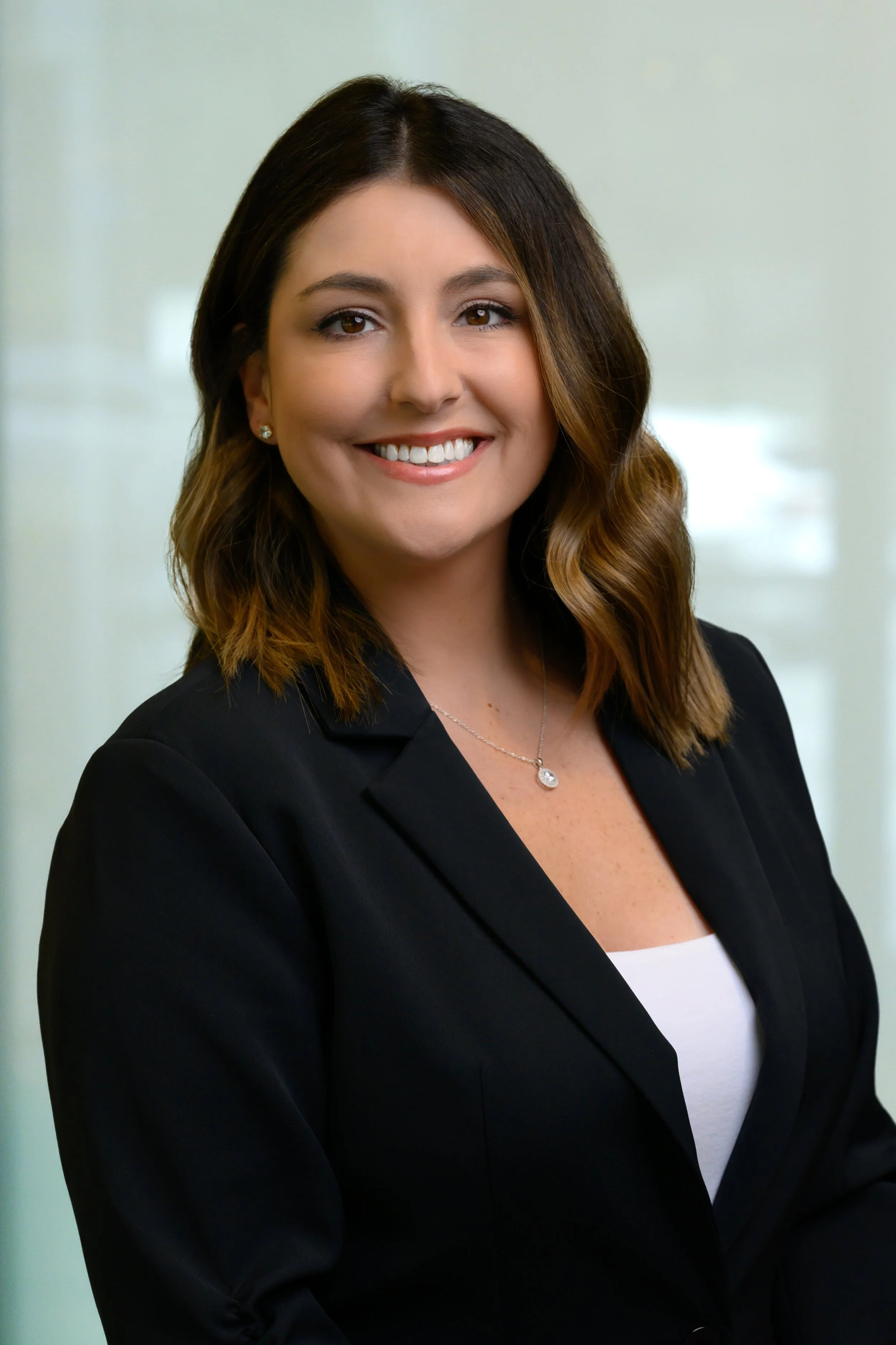 A woman with shoulder-length wavy brown hair smiling, wearing a black blazer, white top, and a necklace with a small pendant, standing in front of a light green background.