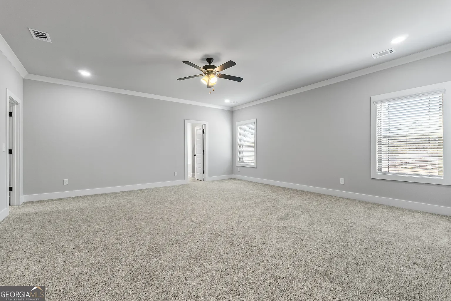 Empty living room with light gray walls, beige carpet, and two windows with blinds. Ceiling fan with lights and a door leading to another room.