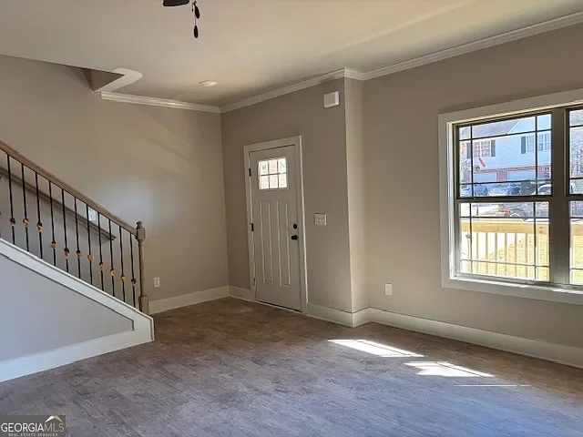 Empty living room with gray walls, a large window showing outdoors, a white front door, a staircase with wooden handrails and black balusters, and hardwood flooring.