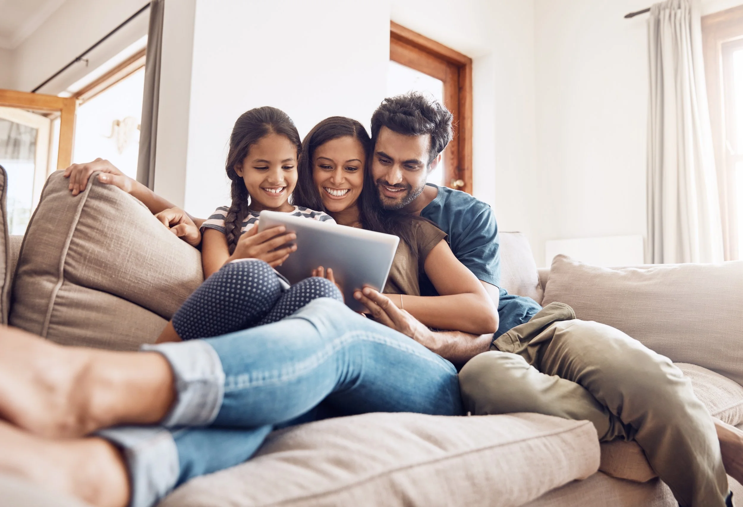 A family of three lounging on a beige sofa, smiling and looking at a tablet together in a bright living room with windows and curtains.